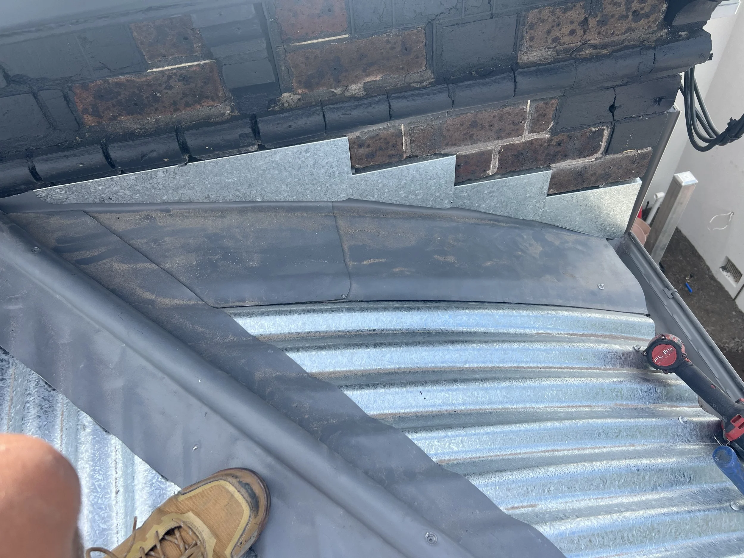 Photo taken from the edge of a roof, showing brown brick chimney with metal flashing. The person's beige work boots are visible in the lower part of the image. A red cordless drill is on the metal roof along with black power cords in the background.