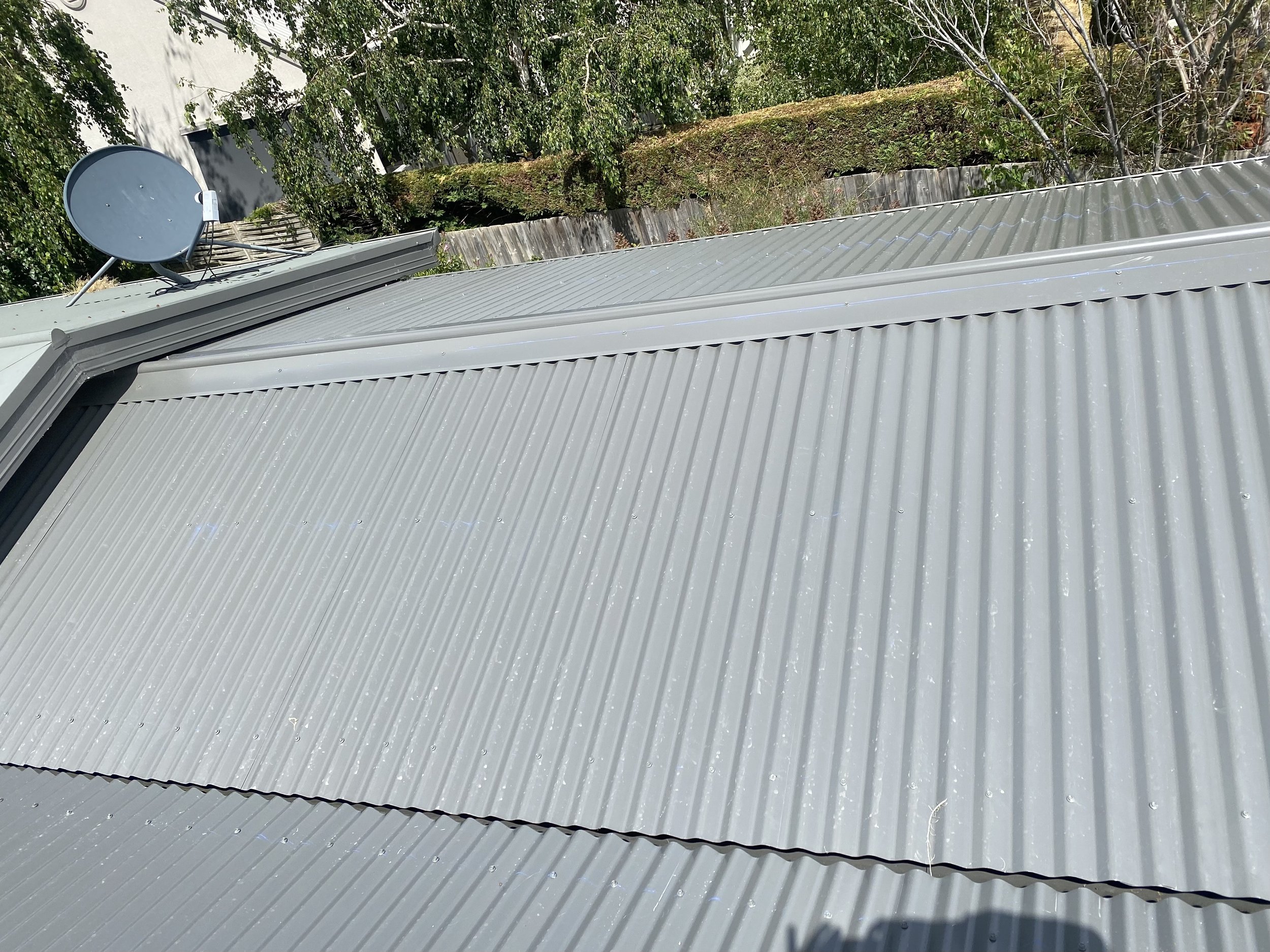 Metal roof with corrugated sheets and a satellite dish mounted on top, with trees and a wooden fence in the background.