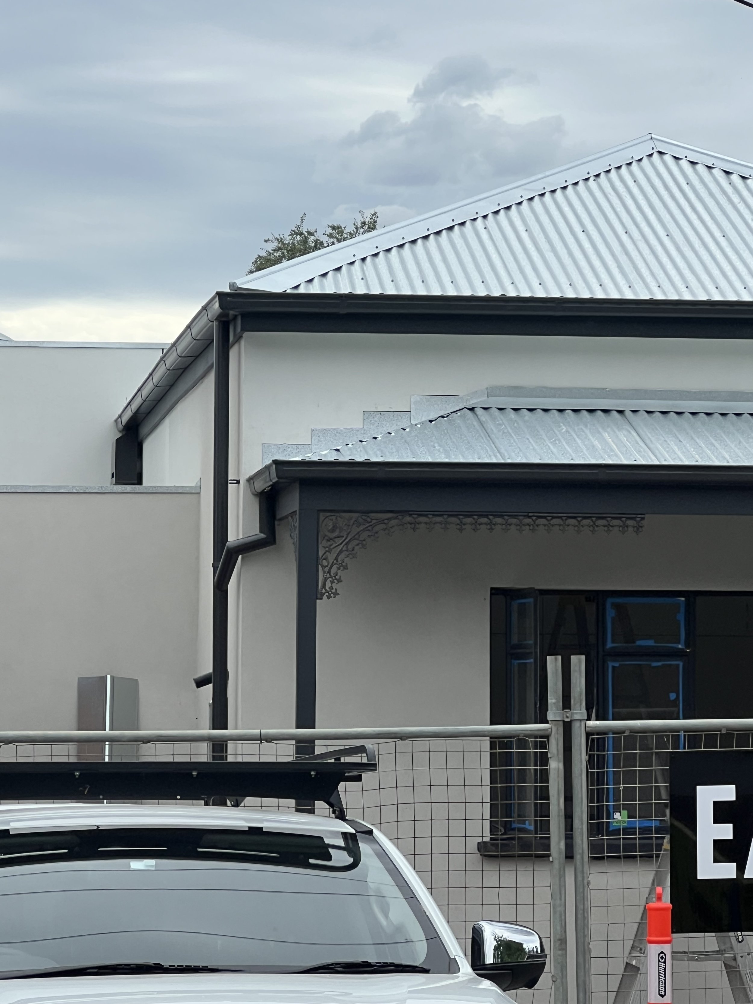 The image shows a modern building with a metallic, corrugated roof and a car parked in front. The building has a black gate and a security fence, with some greenery visible behind the building. There is an overcast sky with clouds.