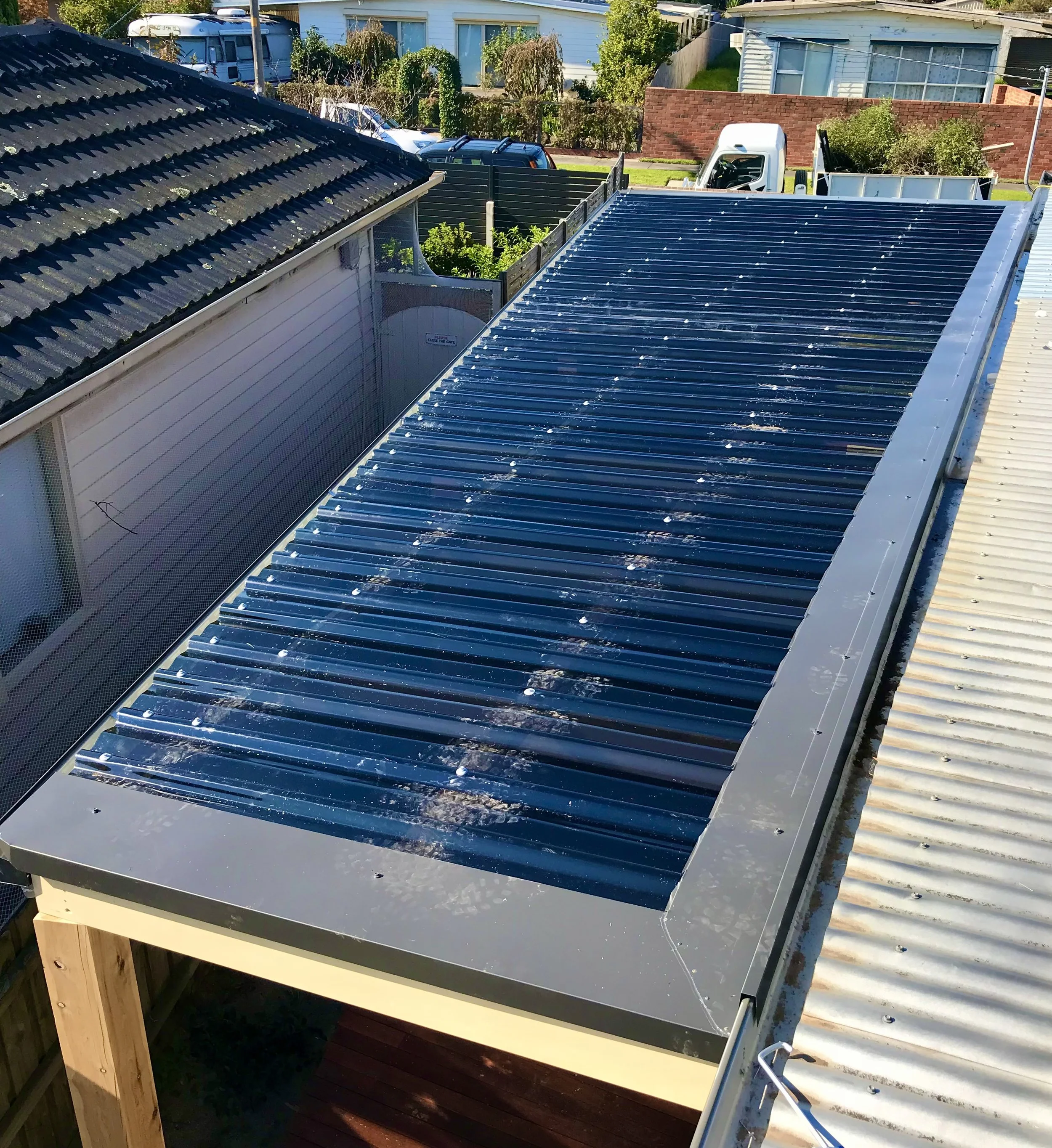 Image of two rooftop solar panel installations, one with black panels on a home roof and another with shiny reflective panels on a nearby metal roof, with backyard fences and houses in the background.