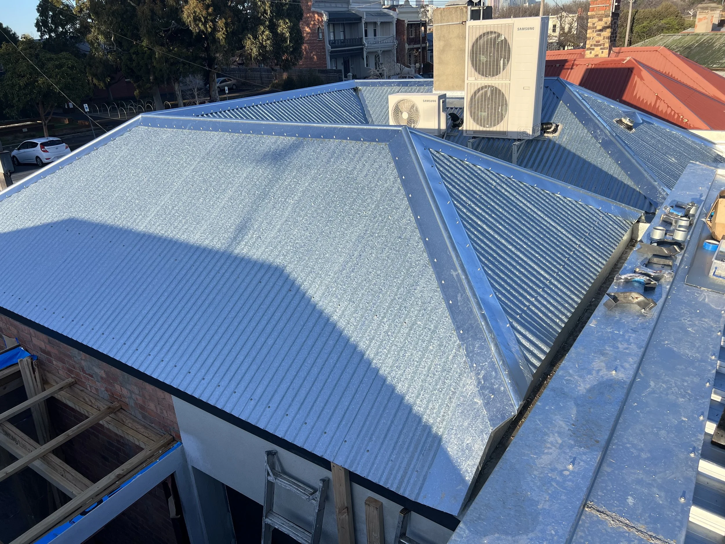 Rooftop view showing a shiny new metal roof with an air conditioning unit, neighboring houses, parked cars, trees, and power lines.