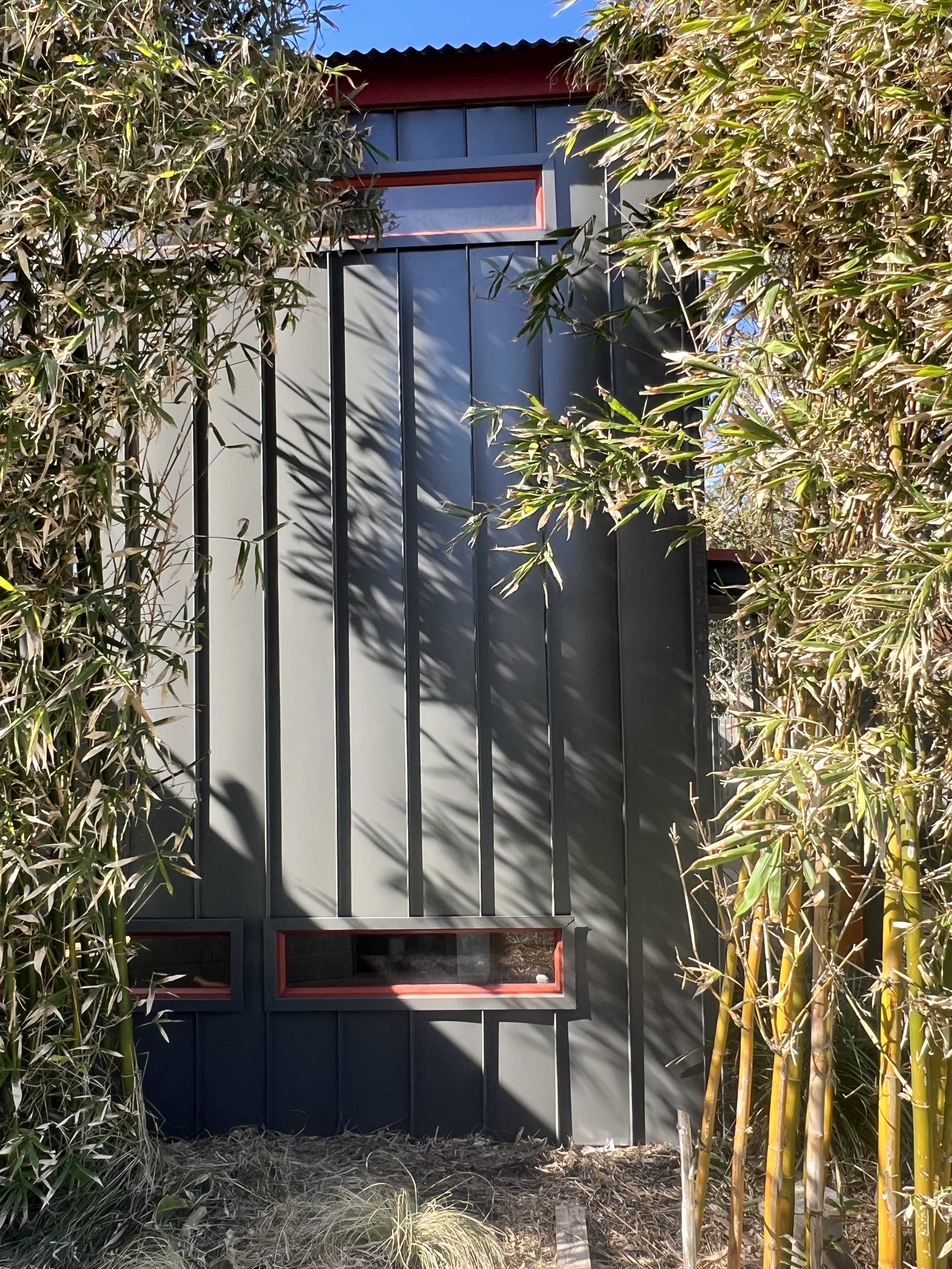 A modern black gate with red accents surrounded by bamboo and shrubs, with a small clearing in front and trees in the background.