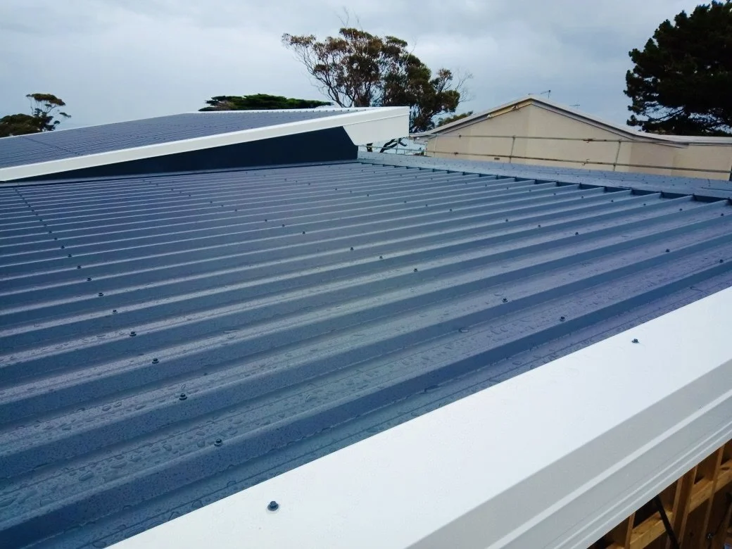 Close-up of a blue metal corrugated roof with raindrops on it, with trees and a house in the background.