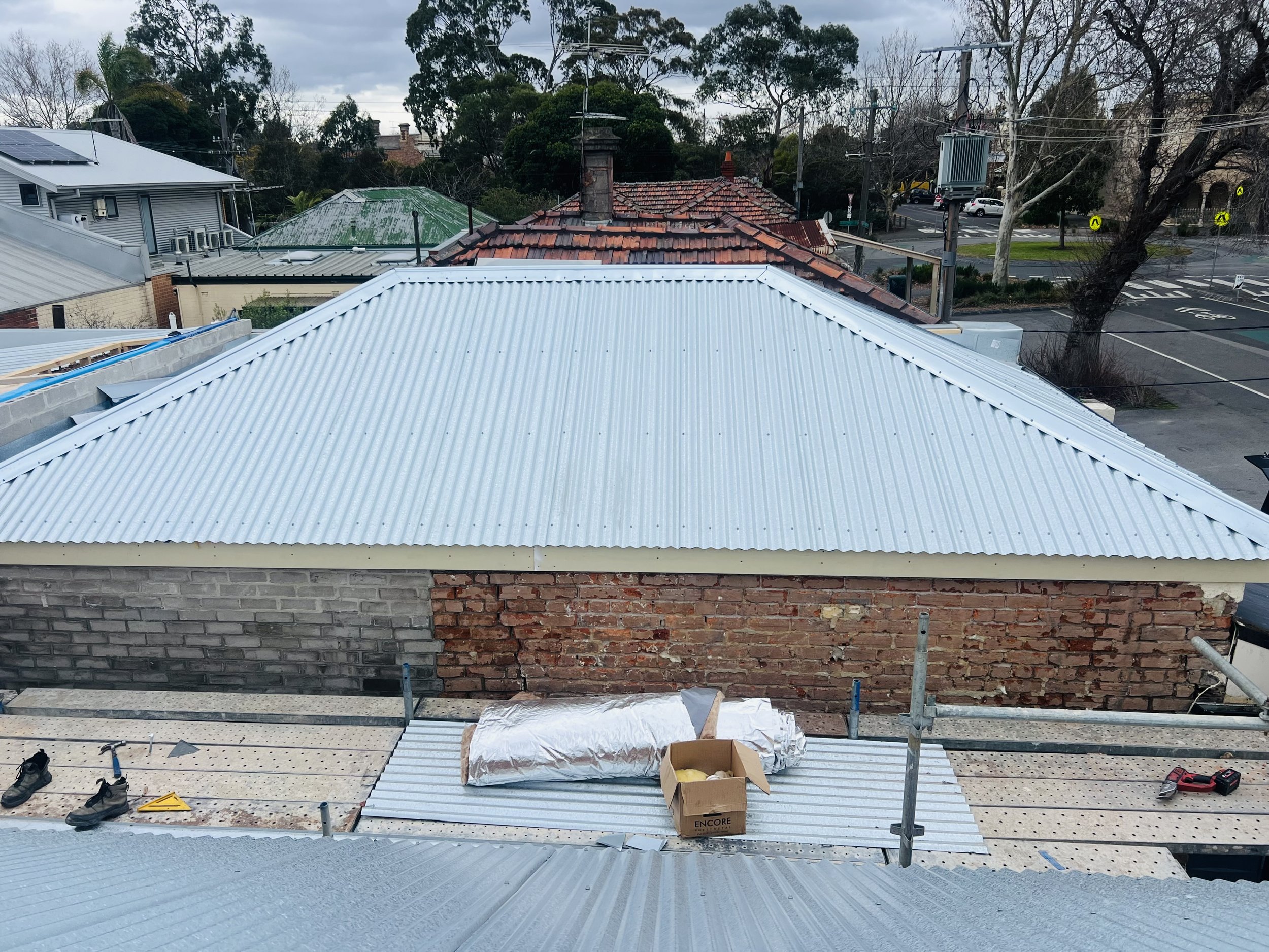 Construction site with a building having a metal roof and brick wall, tools, and materials scattered around; neighboring rooftops, trees, and street with parked cars visible in background.