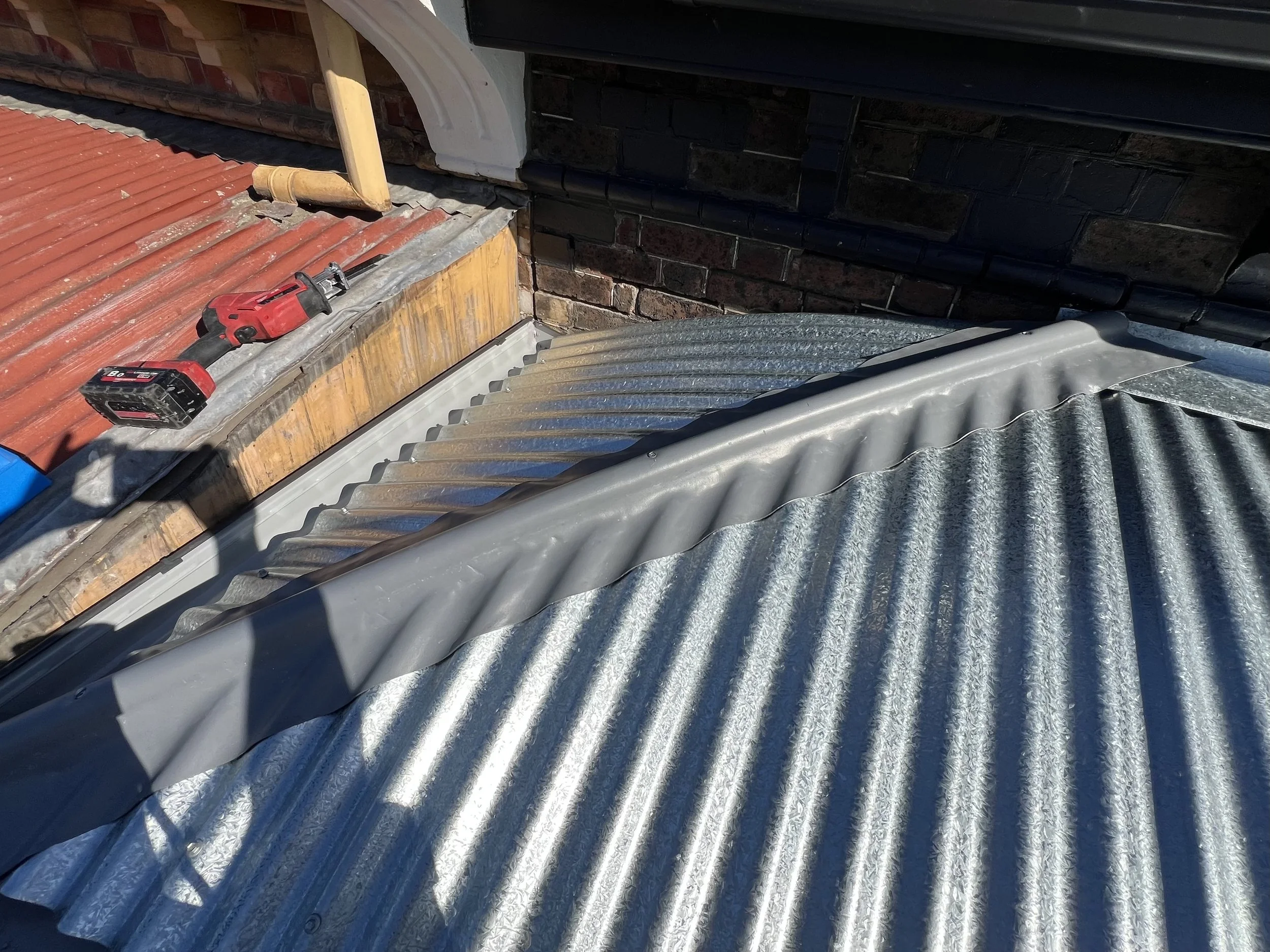 The image shows a rooftop with different materials, including a corrugated metal sheet and a tiled roof. There is a cordless drill lying on the roof, and parts of the building's brick wall and wooden support structures are visible.