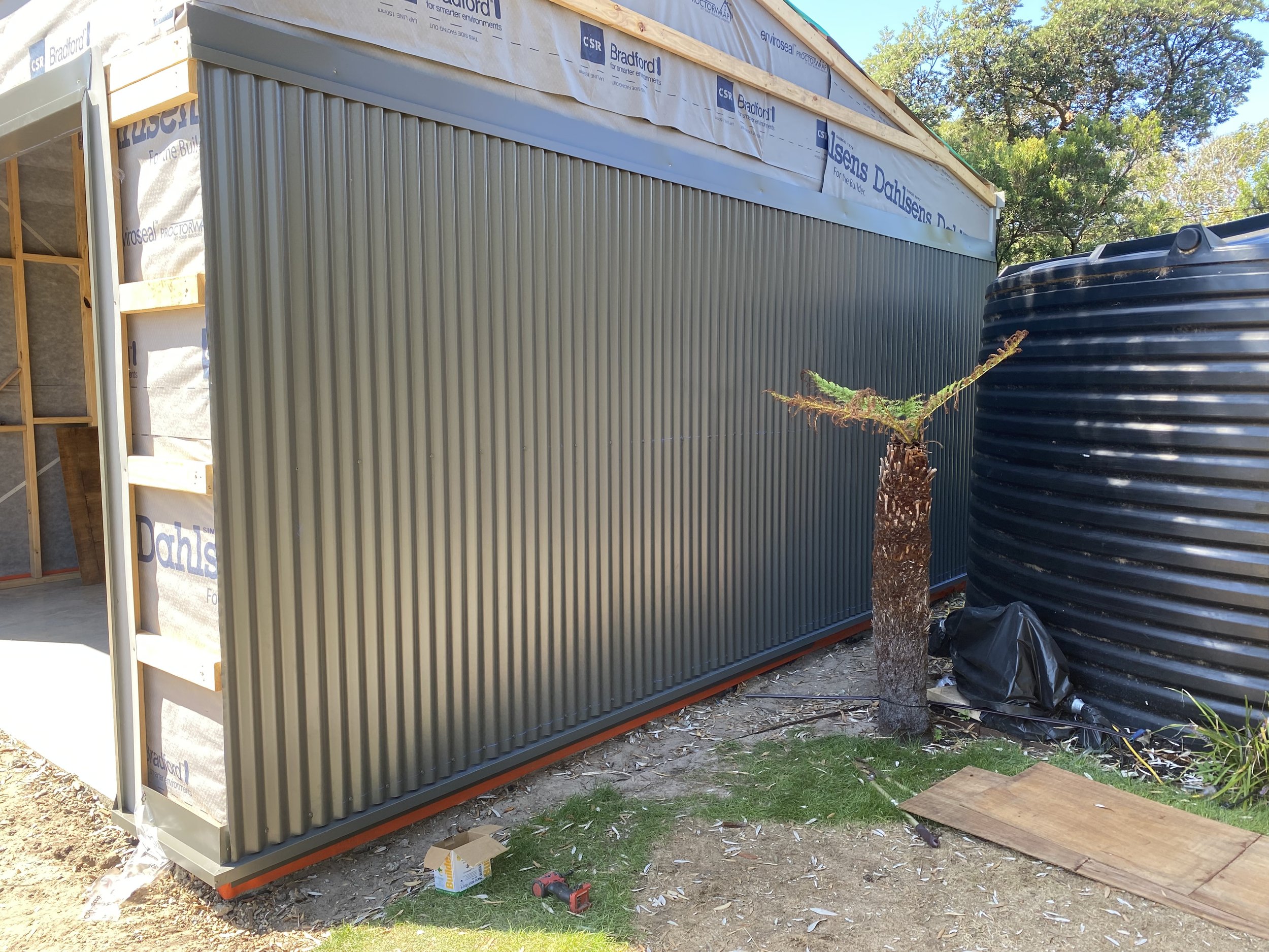 Construction site with a partially built structure featuring metal siding and wood framing. A black water tank and a small tree are visible nearby. The area has construction tools and debris on the ground.