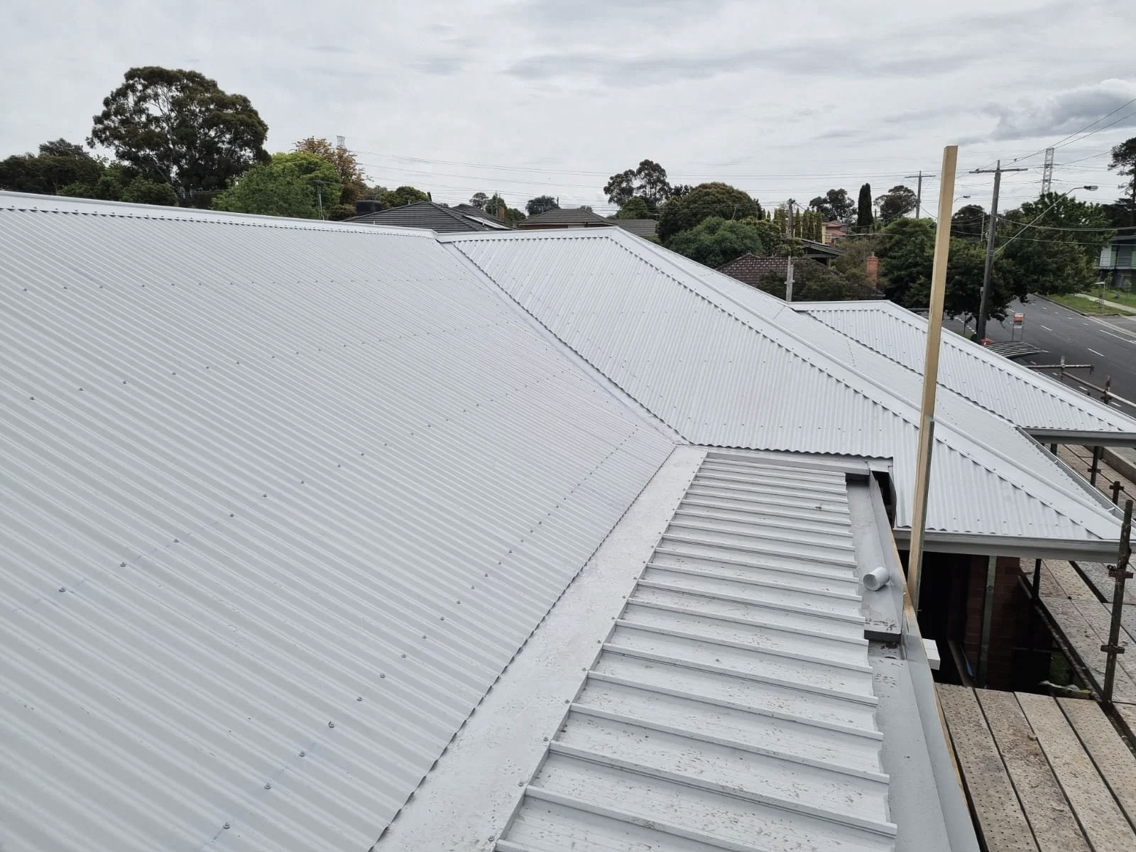 View of a metal roof with multiple sections and a cloudy sky, trees, and some residential houses in the background.