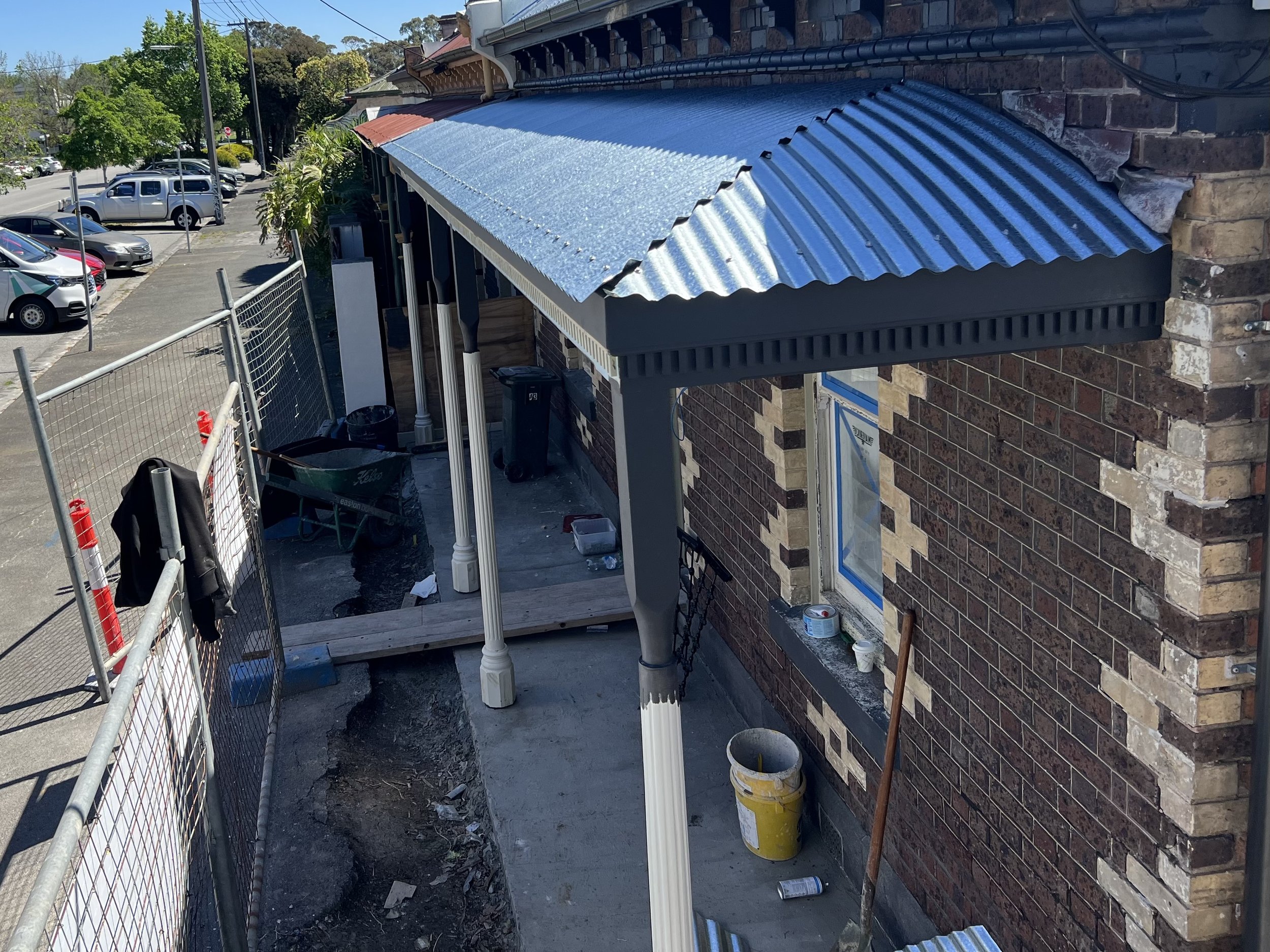Construction area with aluminum roofing panels installed over a small structure attached to a brick building, construction tools and debris on the ground, parked cars and trees in the background.