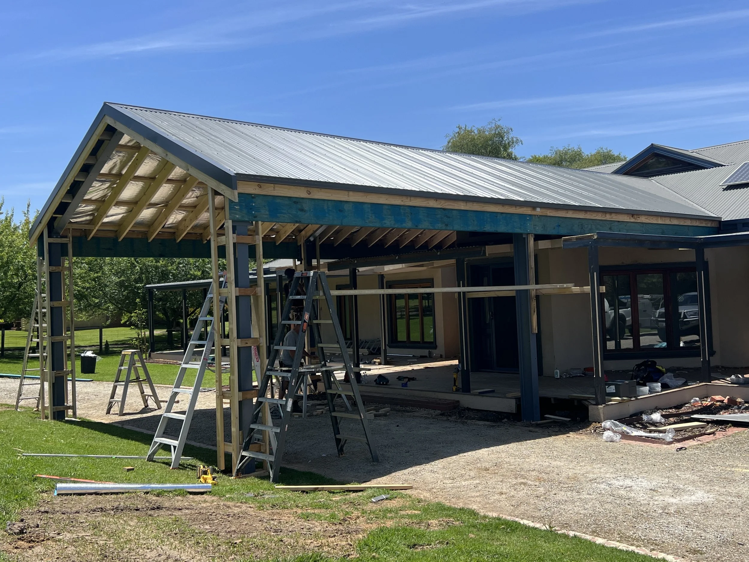 Construction site with a house in progress, showing a partially built roof with metal roofing panels and ladders, under a clear blue sky.