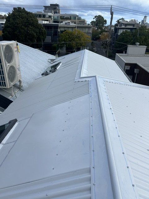 View of a white metal roof with multiple slopes, an air conditioning unit, and air vents, with a cityscape and trees in the background.