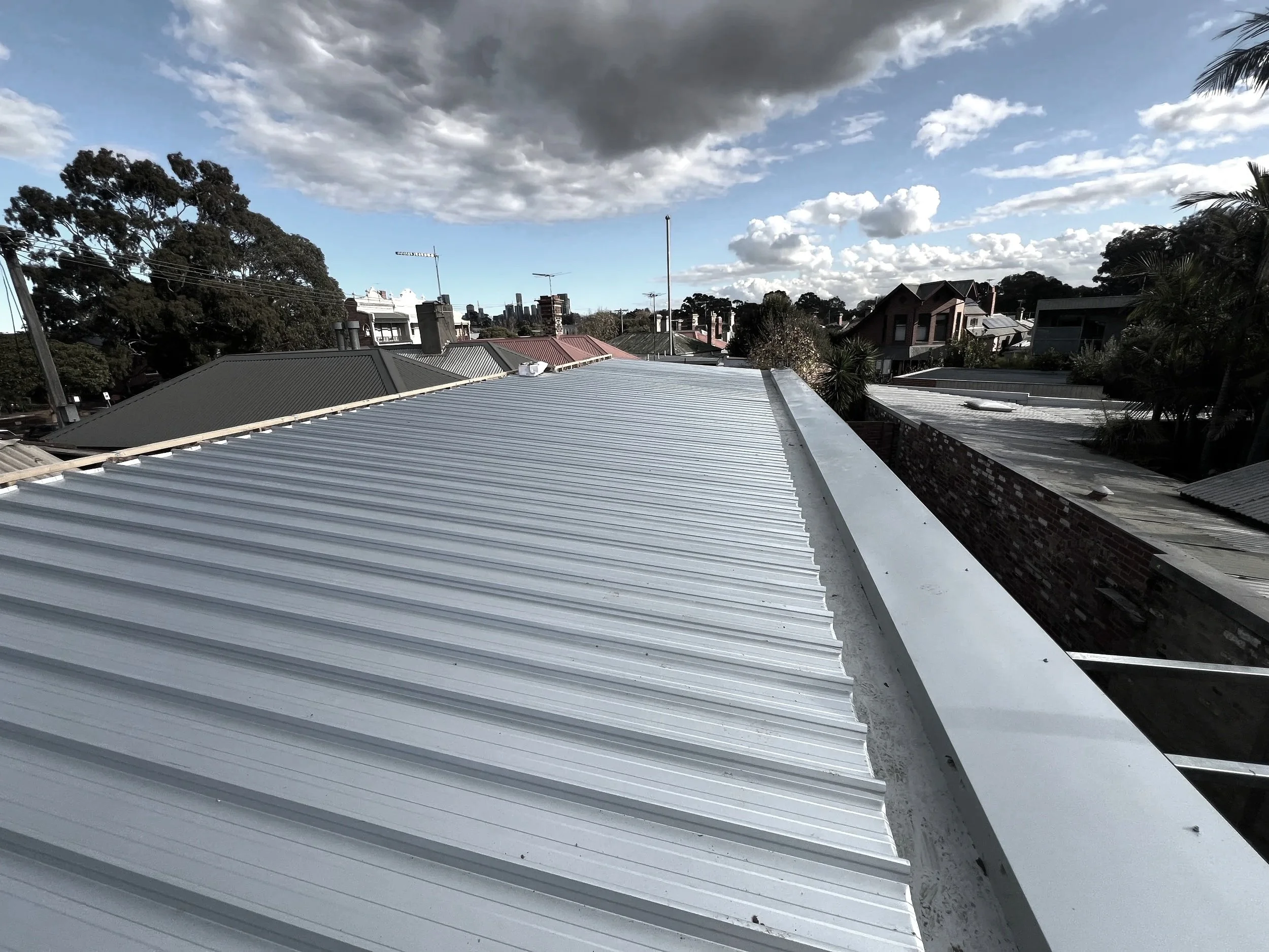 View of rooftops with metal and tiled roofing, trees, and a cloudy sky over a residential area.