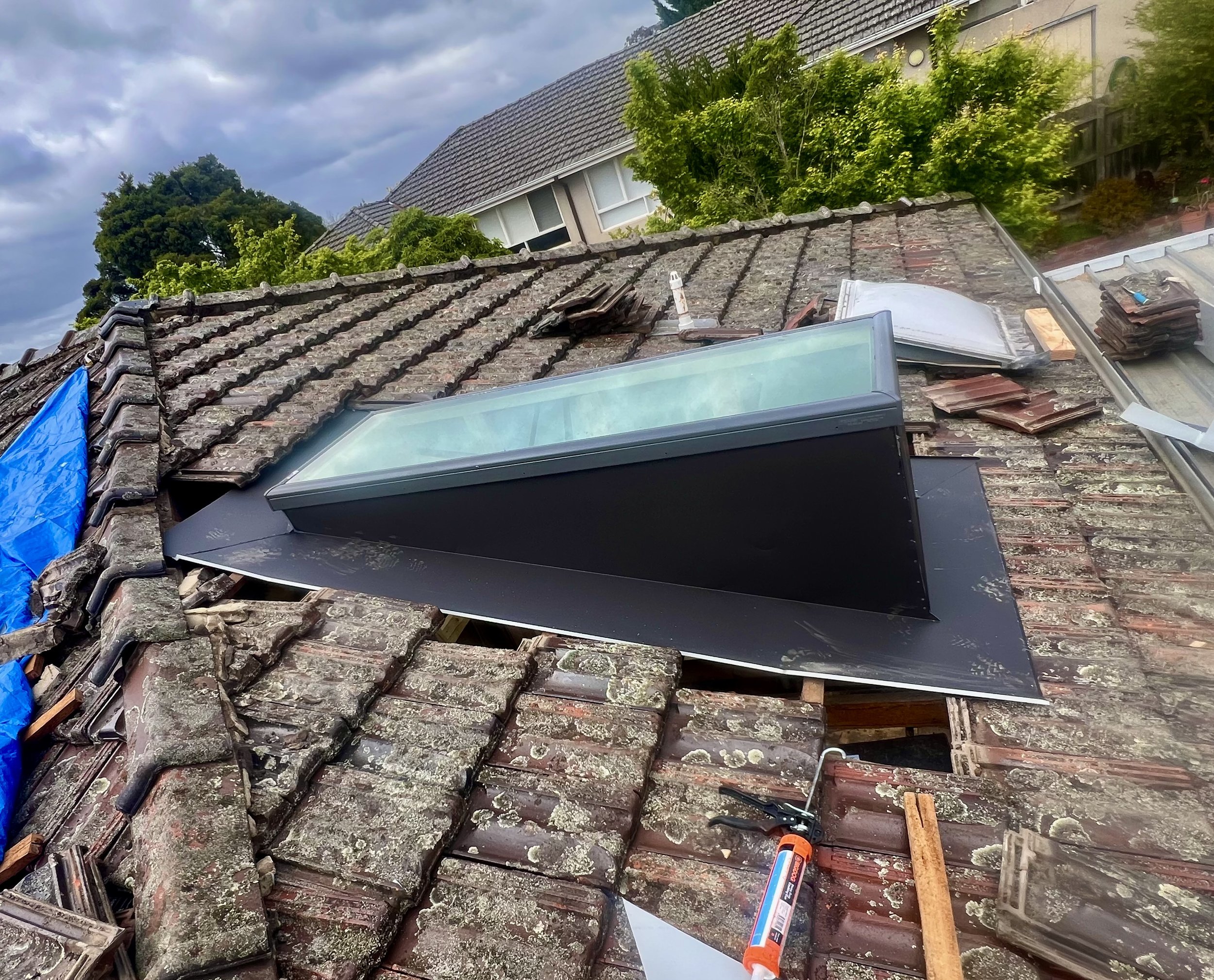 View of a rooftop with old tiles and debris, tools, and roofing materials, with trees and a house in the background under a cloudy sky.