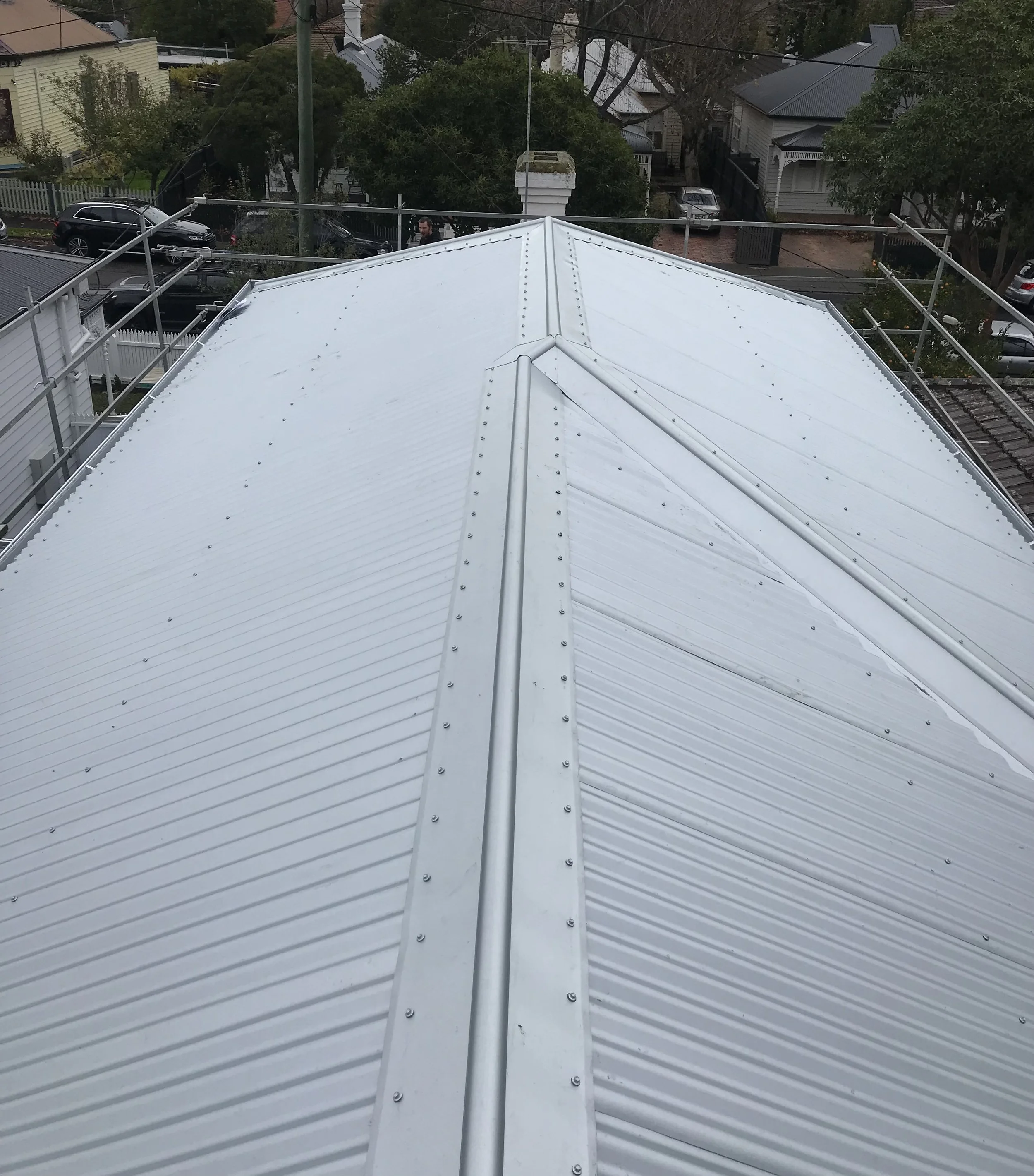 View from the top of a building showing a white metal roof with visibility of neighboring houses, trees, parked cars, and a person walking on the street below.