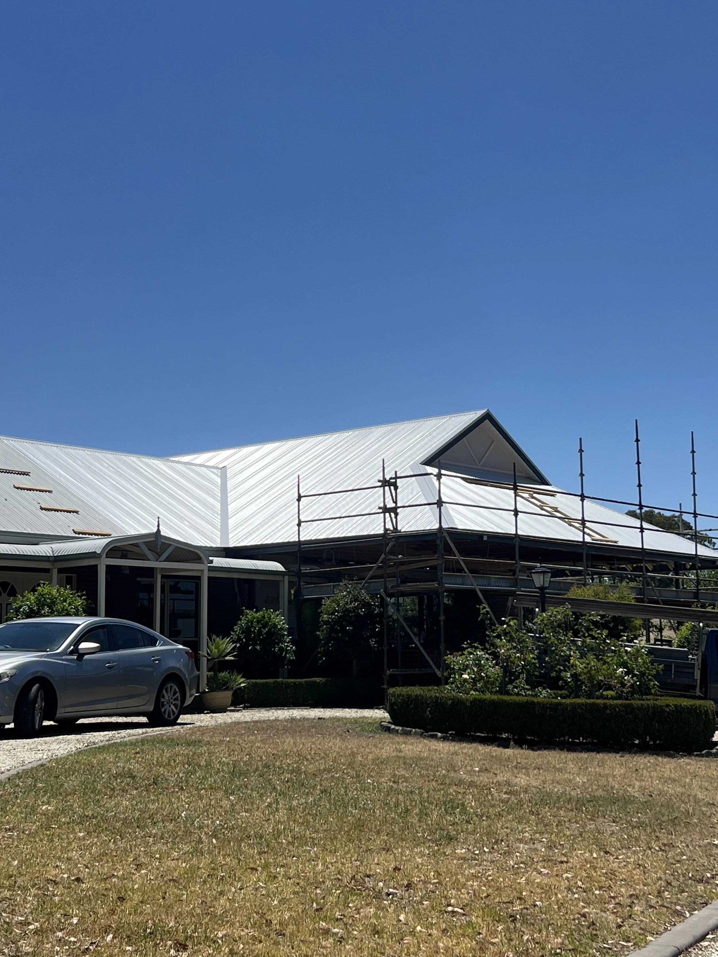 Building under construction with white metal roof and scaffolding, parked gray car in front, grassy area with patches of dirt and small bushes, clear blue sky in the background.