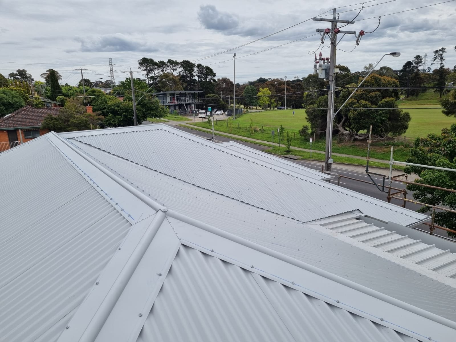 View of a white corrugated metal roof on a building, with a grassy park, trees, power lines, and a cloudy sky in the background.