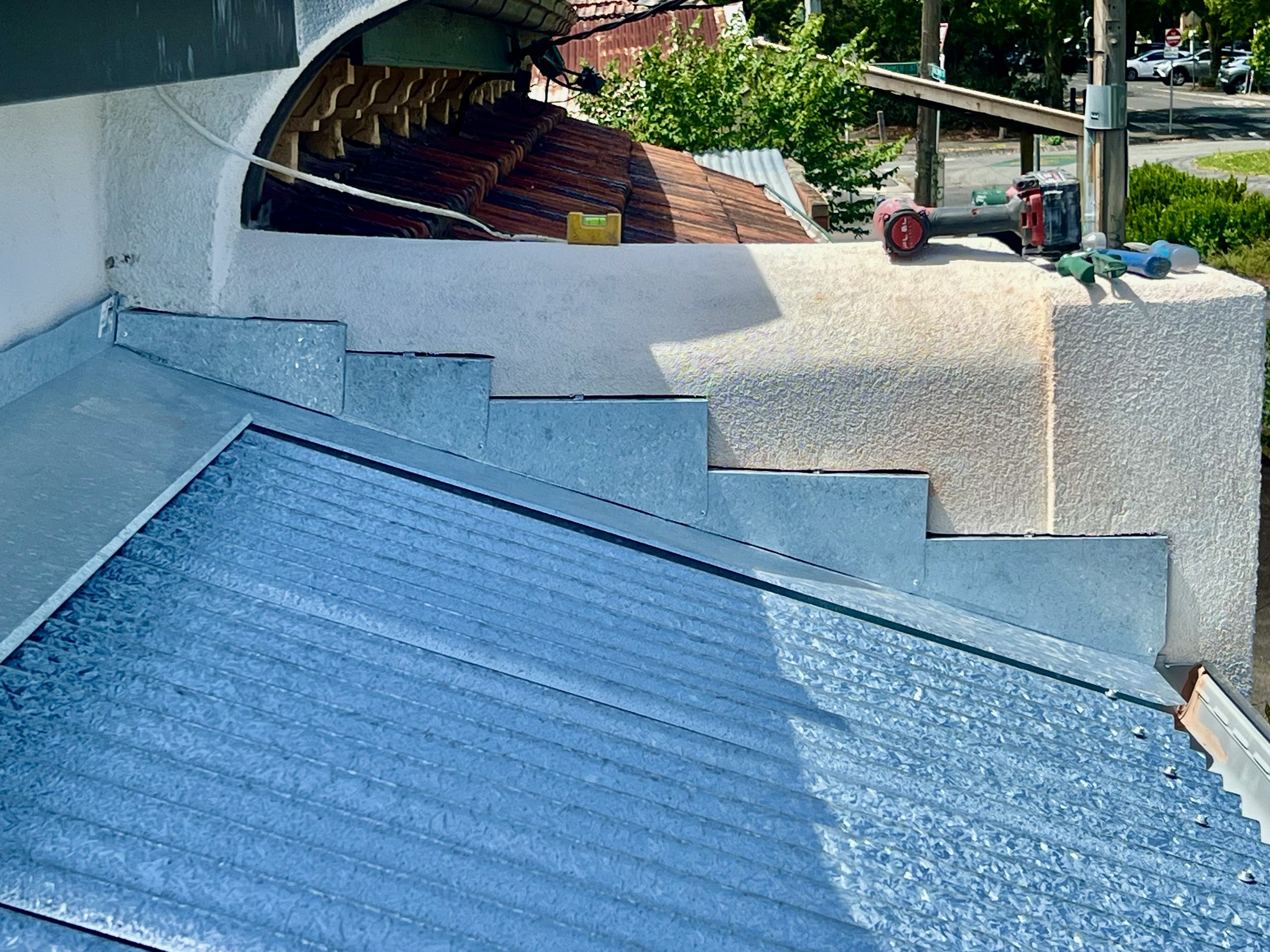 View of a building's rooftop with shingles, some electrical wiring, a small yellow toolbox, and a cordless drill resting on the edge. In the background, trees, a street with parked cars, and utility poles are visible.