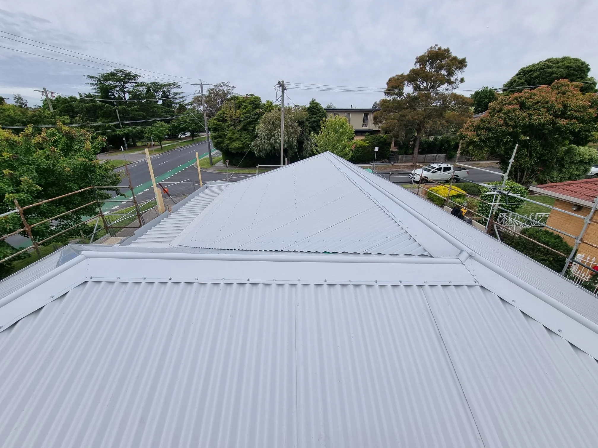 View from a rooftop showing a suburban street with trees, cars, power lines, and cloudy sky.
