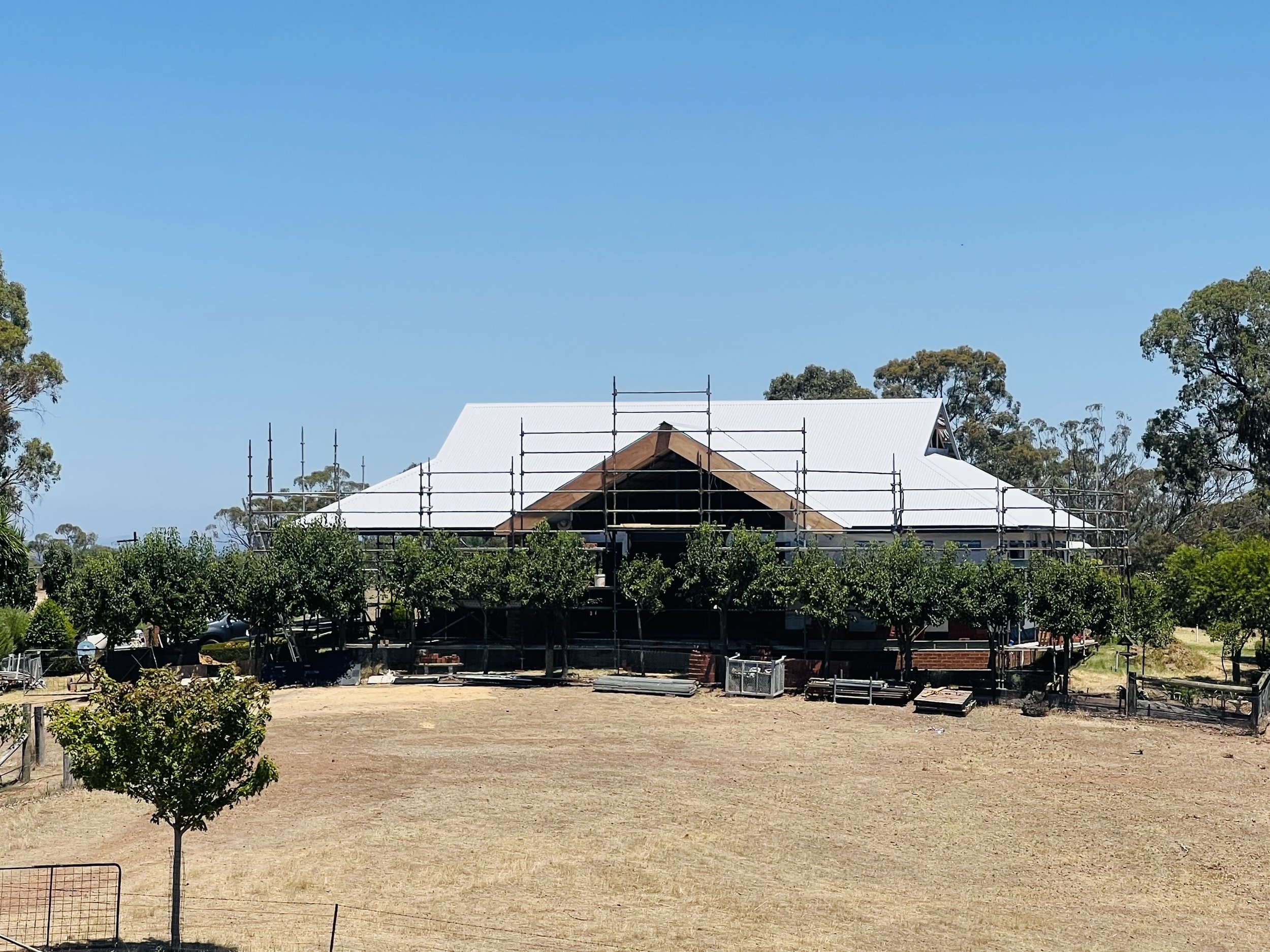 A house under construction with a white roof, surrounded by scaffolding, in a rural setting with trees and open land.