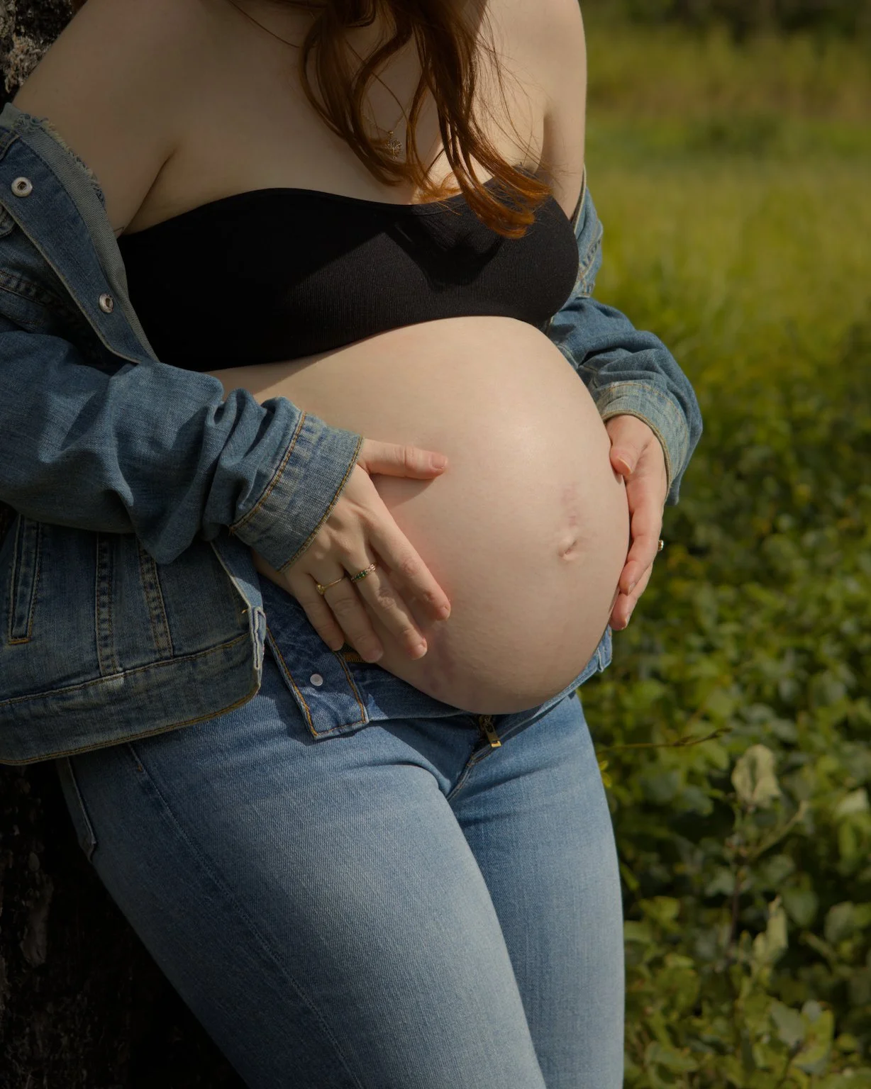 A pregnant woman with red hair holding her pregnant belly outdoors, wearing a black top, an unbuttoned denim jacket, and blue jeans, with greenery in the background.