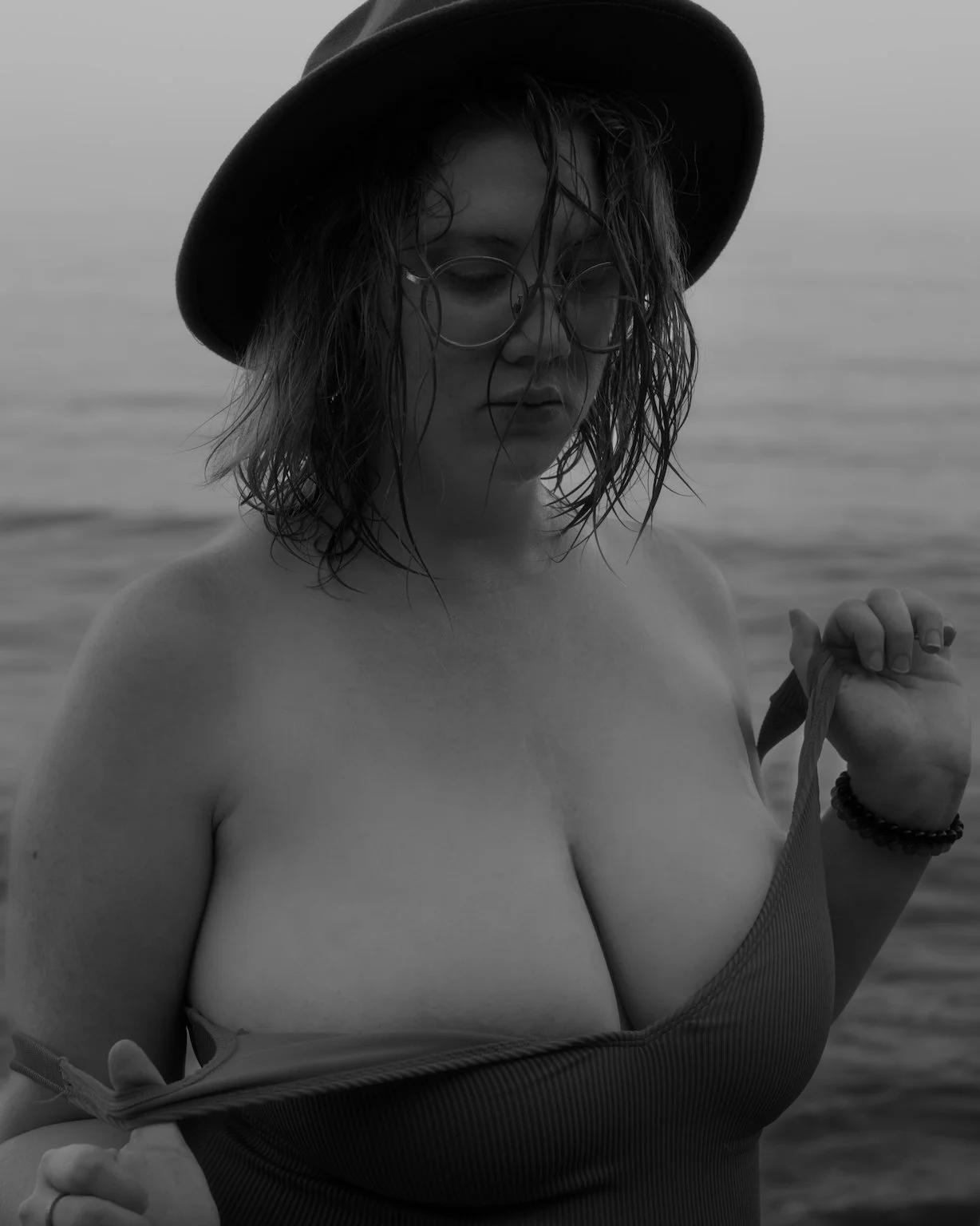 Black and white photo of a vintage car driving down a rural road. The road is flanked by grass and surrounded by hills with a light cloud cover. There are bird silhouettes flying in the air and utility poles lining the road.