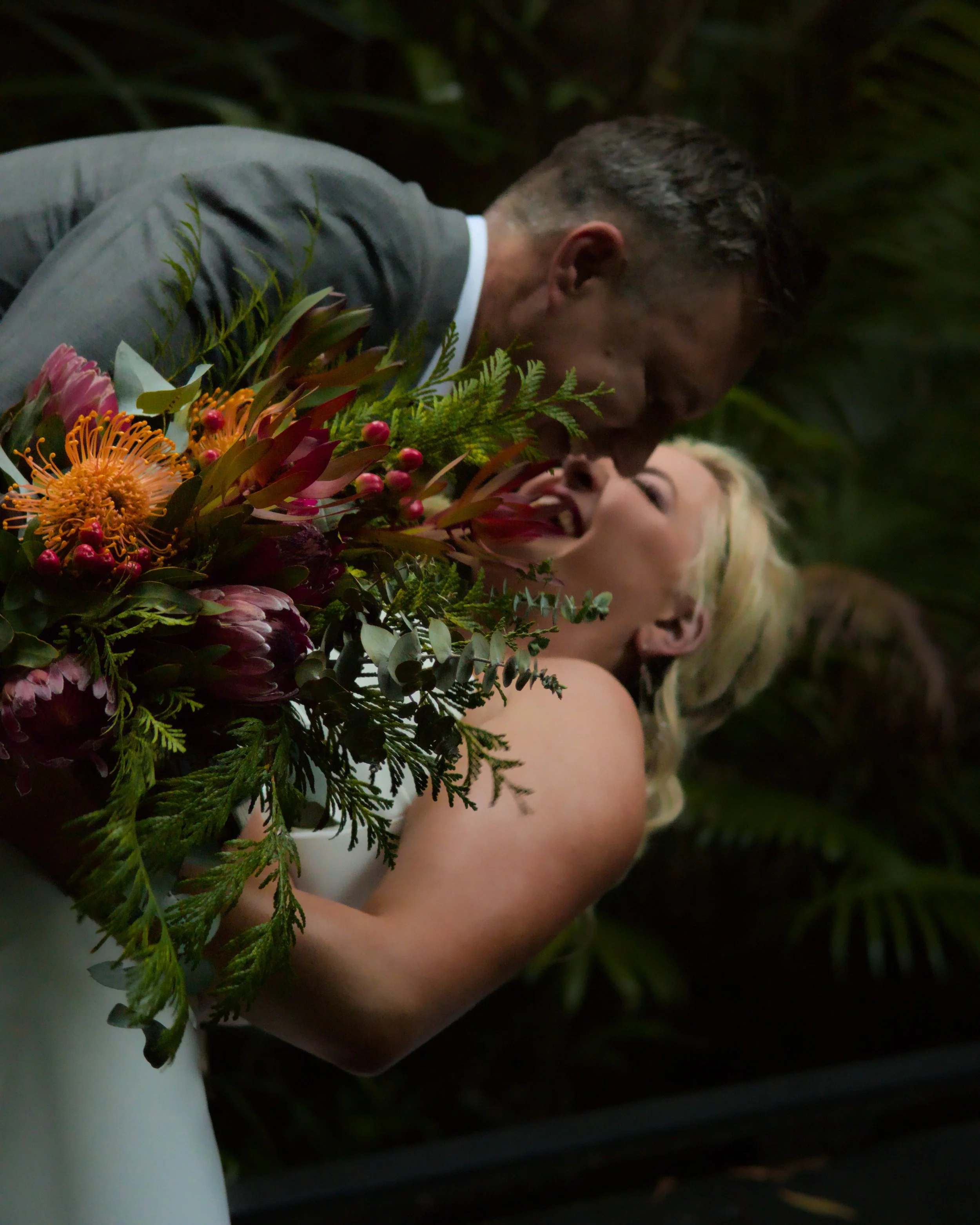 A couple is kissing at their wedding, with the bride holding a large bouquet of colorful flowers.