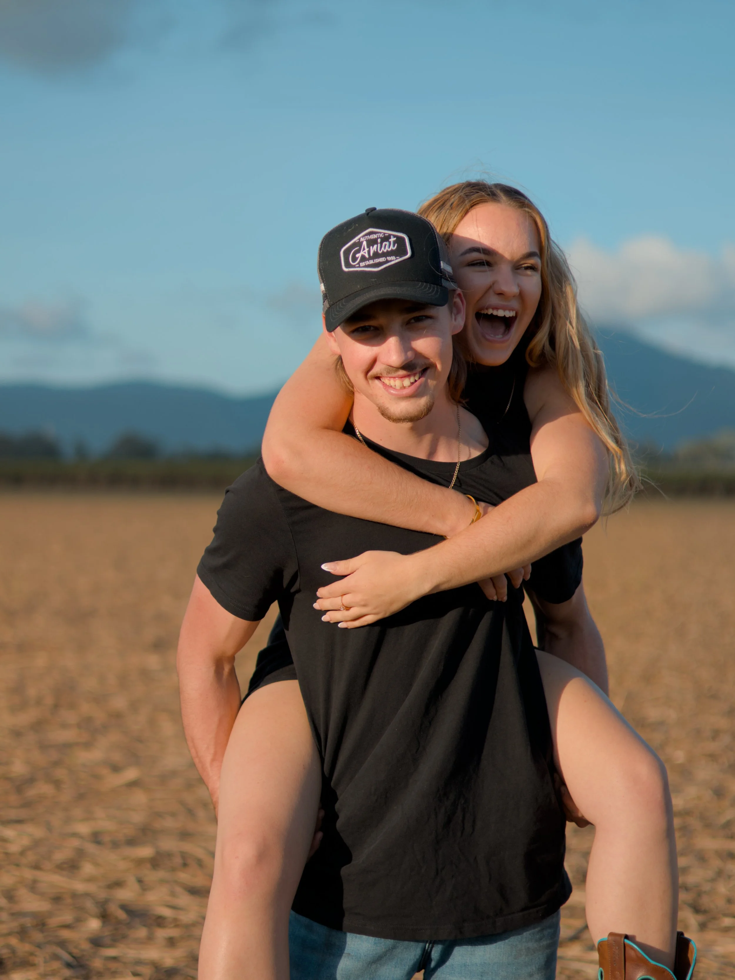 A young man giving a piggyback ride to a young woman in an open field with mountains in the background, both smiling and enjoying the moment.