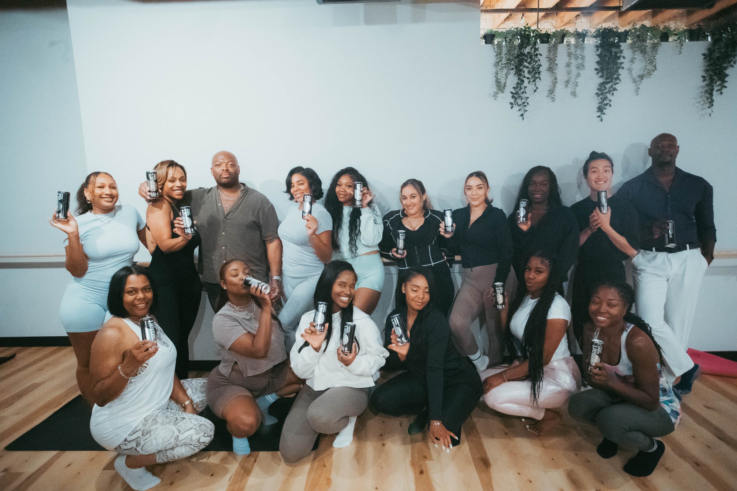Group of diverse women and two men gathered indoors, holding cans of beverage, smiling for the camera in a fitness or wellness class setting.