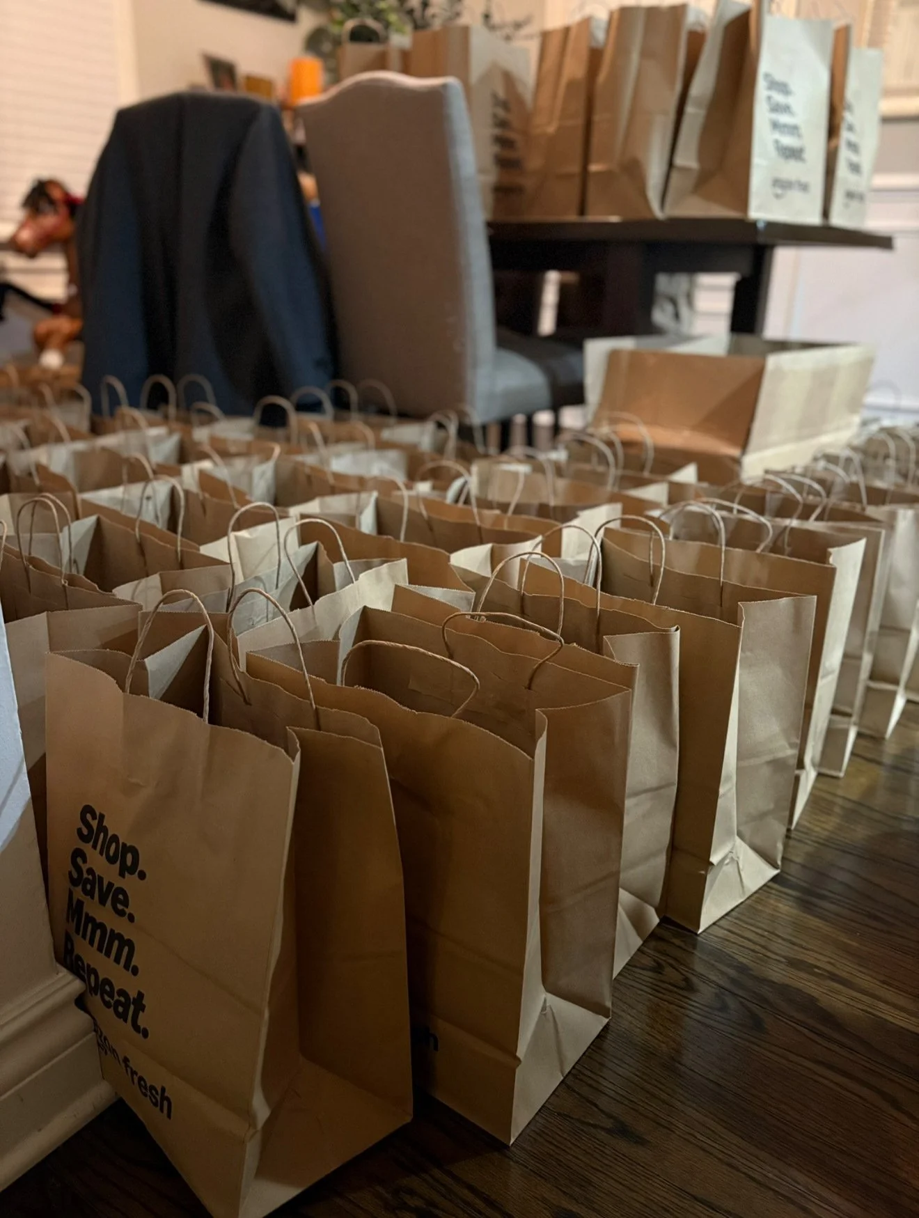 Multiple brown paper grocery bags lined up on a wooden floor, with a table and chairs in the background.
