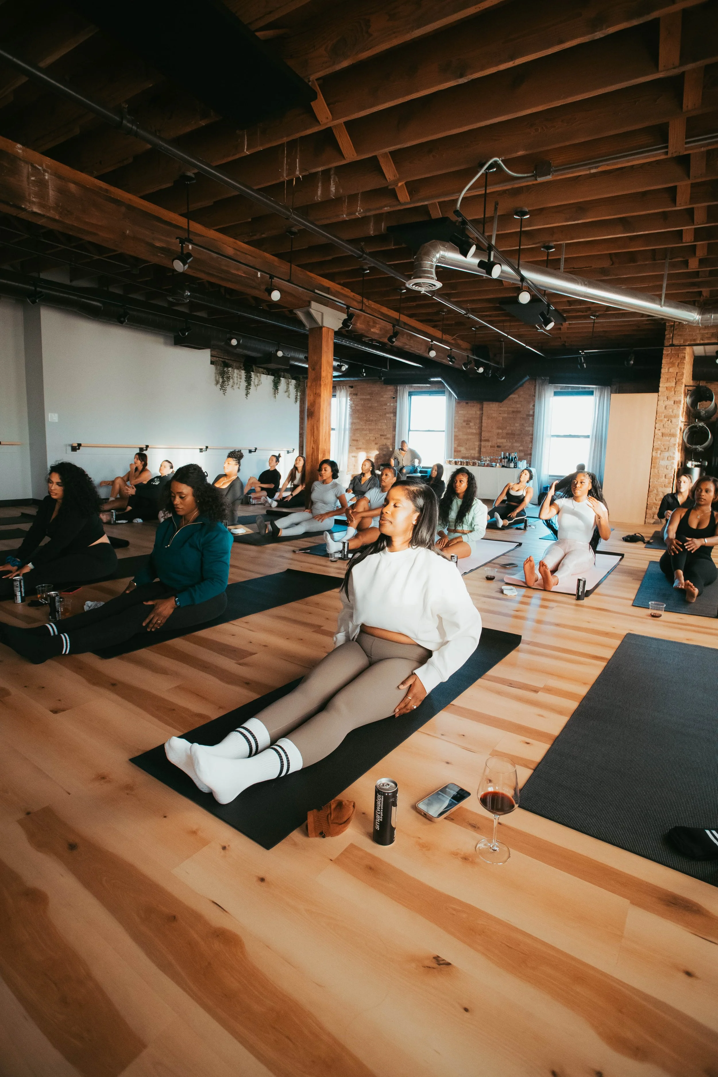 A group of women practicing yoga or meditation in a spacious, wooden-floored studio with natural light coming from large windows.