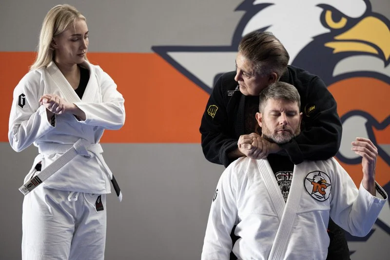 A female Brazilian Jiu-Jitsu practitioner in a white gi and a male in a white gi with an eagle logo, receiving a demonstration from a male instructor in a black gi with yellow patches in a martial arts training facility.