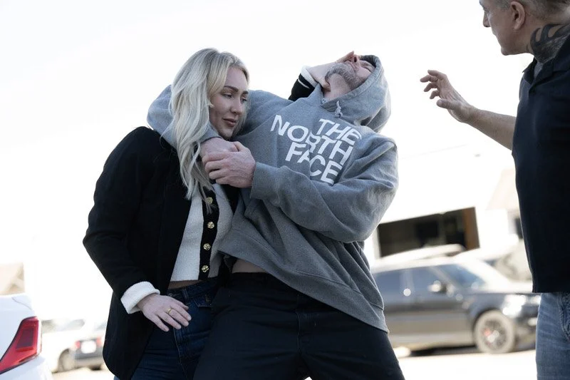 A woman with blonde hair in a black jacket standing next to a man in a gray hoodie with The North Face logo, who appears distressed, while a man on the right gestures with his hand.