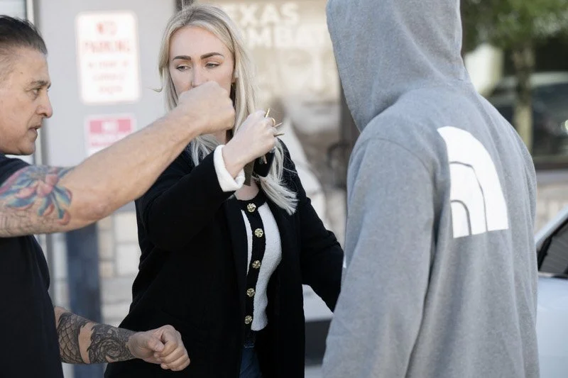 Three people are standing outdoors, talking. The woman in the middle has blonde hair, skin, and is wearing a black jacket with gold buttons, holding her hands up. The man on her left has tattoos on his arm and is reaching out, possibly adjusting the 