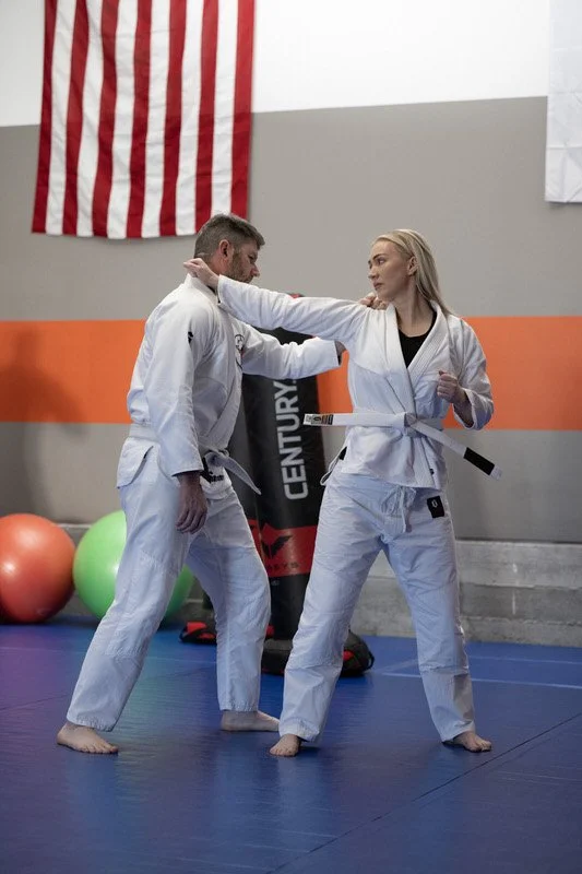 Two women practicing martial arts in a gym, one helping the other with a fighting stance, both wearing white gis and belts.