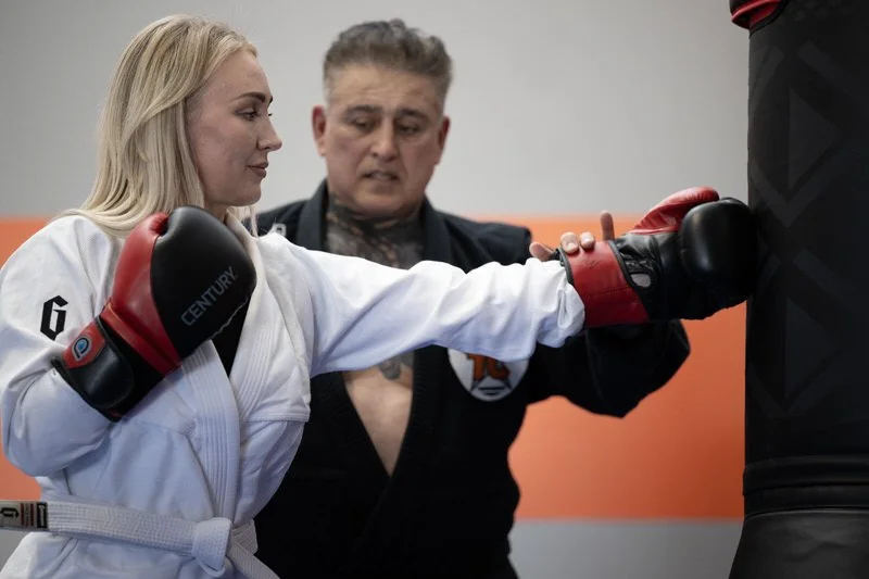 A female martial artist practicing punches on a punching bag, supervised by an instructor in a martial arts gym.