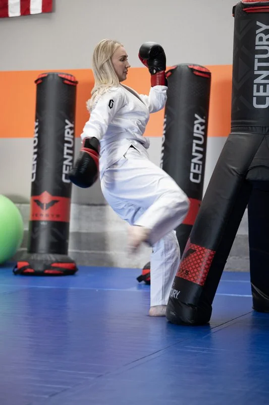 A woman practicing martial arts in a dojo, wearing a white gi and black and red gloves, surrounded by punching bags.