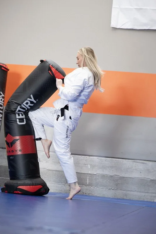 A young girl practicing martial arts on a blue mat, delivering a kick to a black and red punching bag in a training gym.