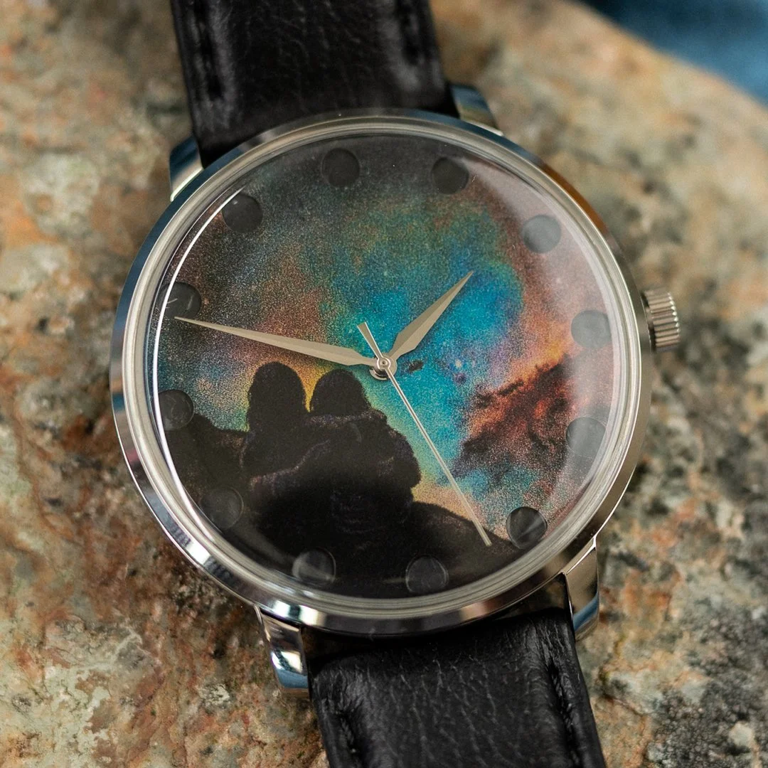 A wristwatch with a space-themed galaxy design on the face, black leather strap, silver casing, and clear glass cover.