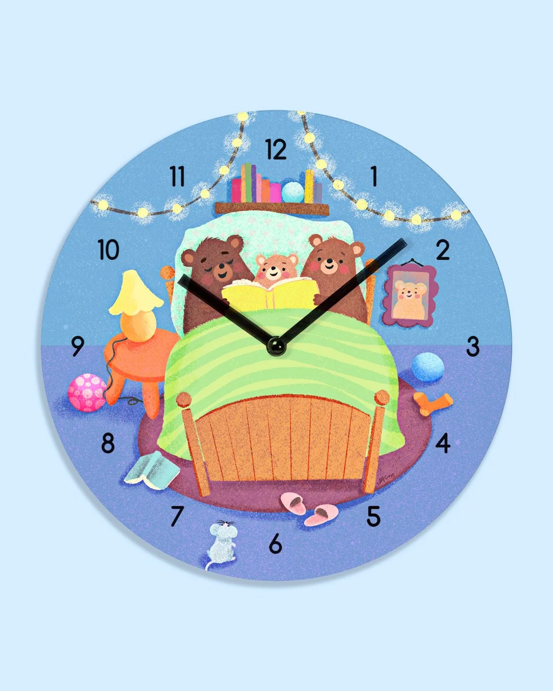 Colorful clock featuring three bears reading in bed, a nightstand with a lamp, framed picture, books, and decorative balls, set against a background with string lights.