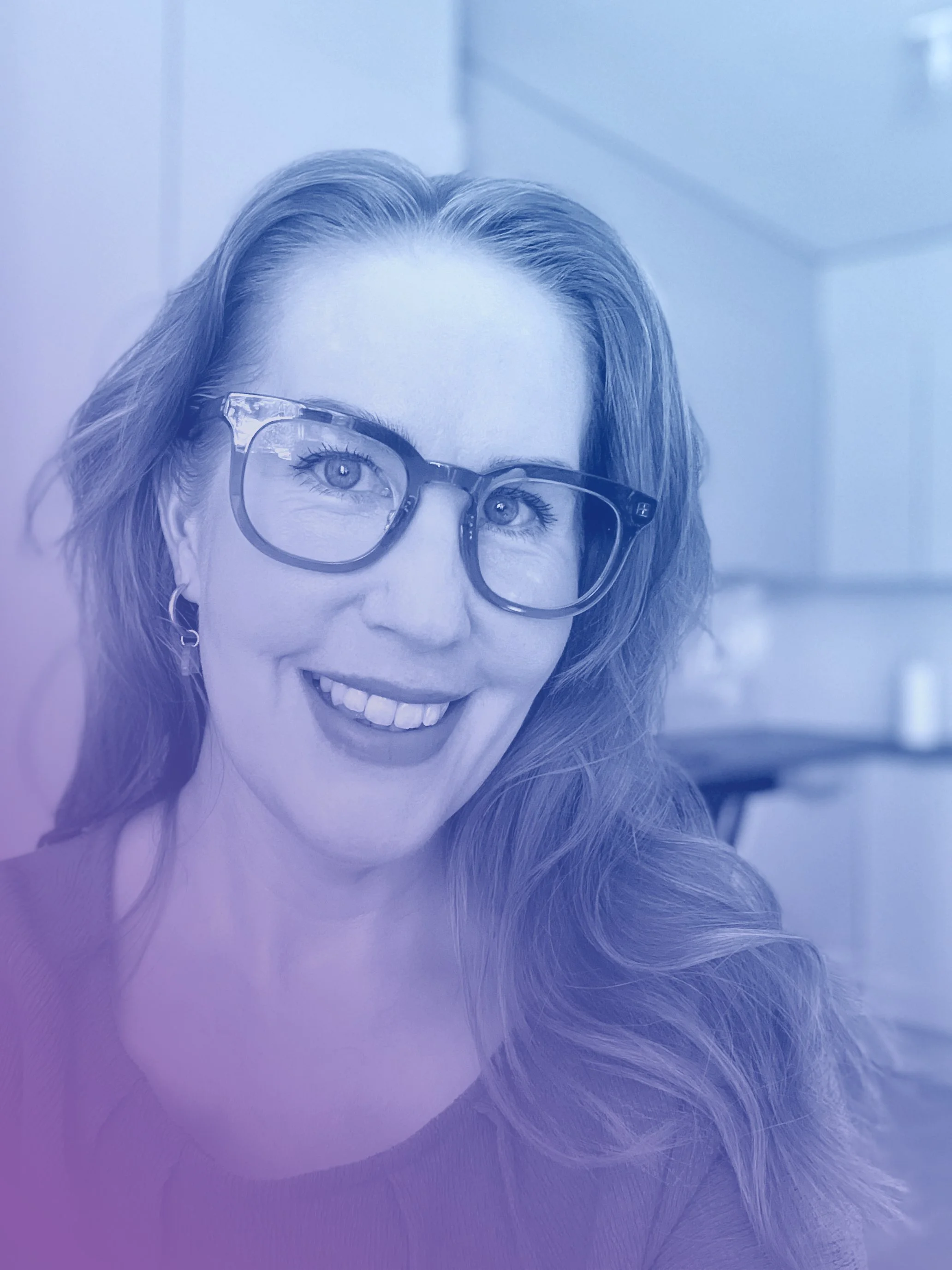 Close-up of a woman with glasses smiling, with brown hair, in a brightly lit room.