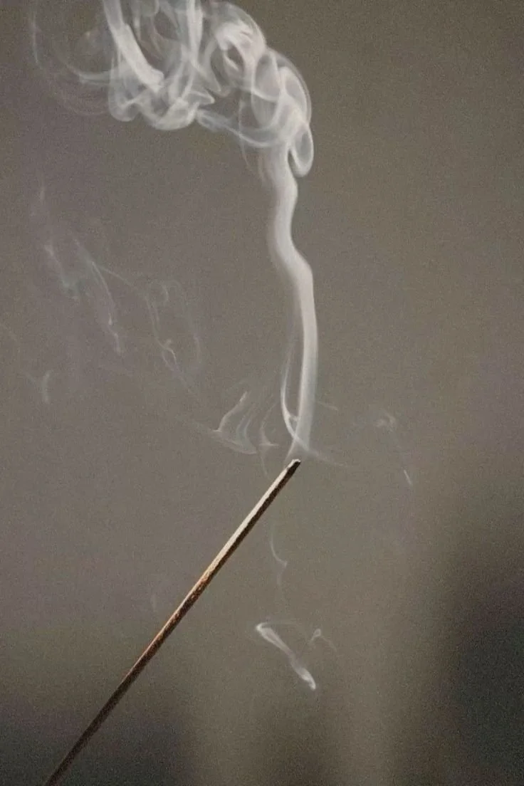 A lit incense matchstick releasing swirling smoke against a plain background.