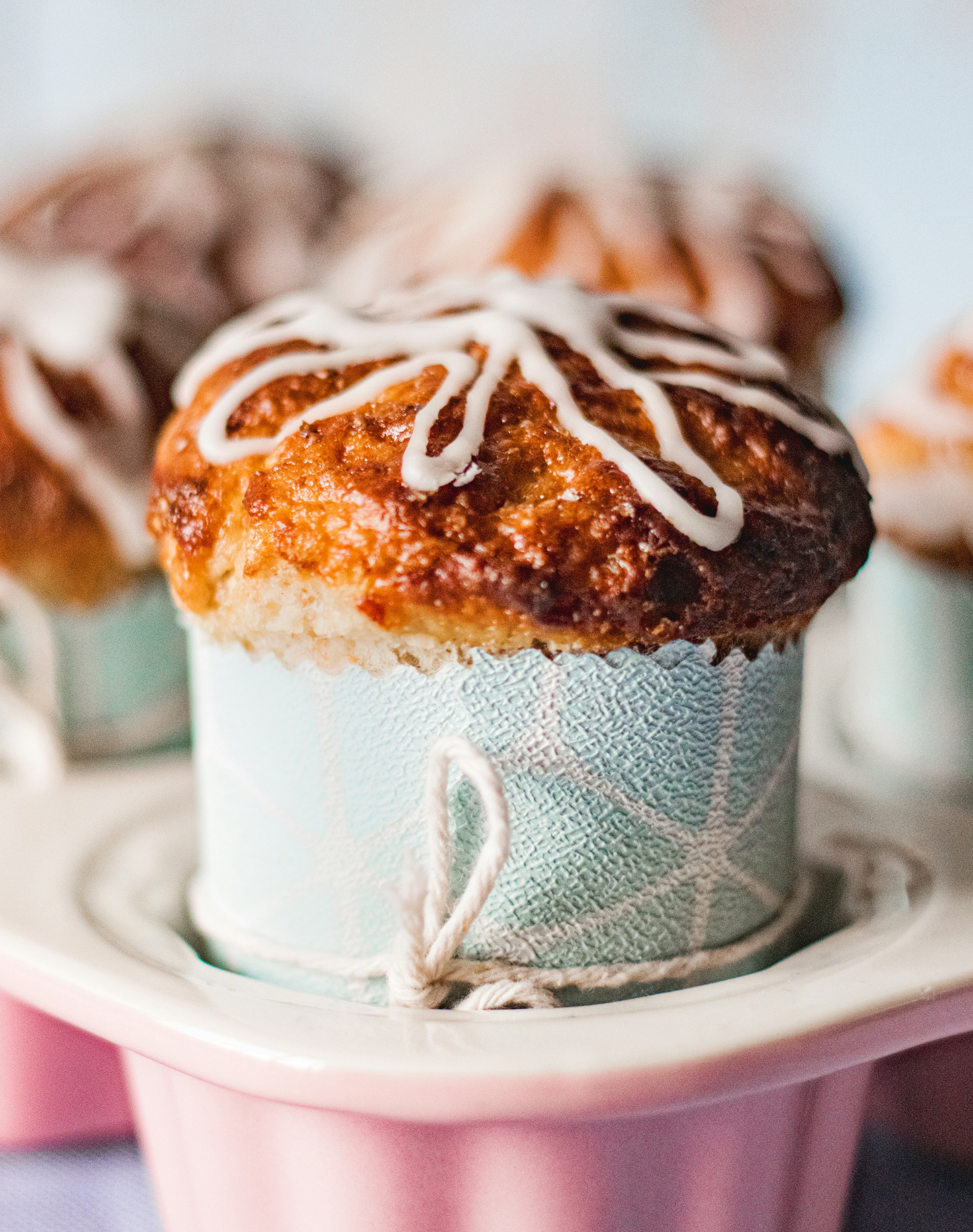 Close-up of a chocolate chip muffin with white icing on top, in a decorative paper wrapper, on a pink plate.