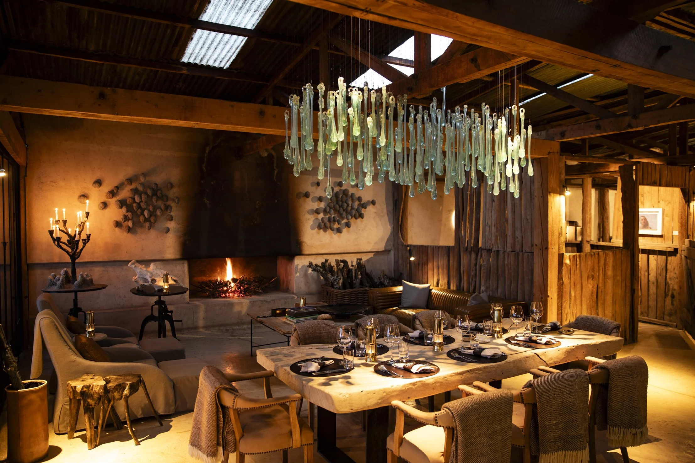 A cozy rustic dining room with a large wooden table set with plates, glasses, and napkins, surrounded by various chairs and couches. A modern chandelier hangs above the table, and there's a fireplace on the back wall. The room features wood paneling, a candelabra with lit candles, and some wall decorations, creating a warm and inviting atmosphere.