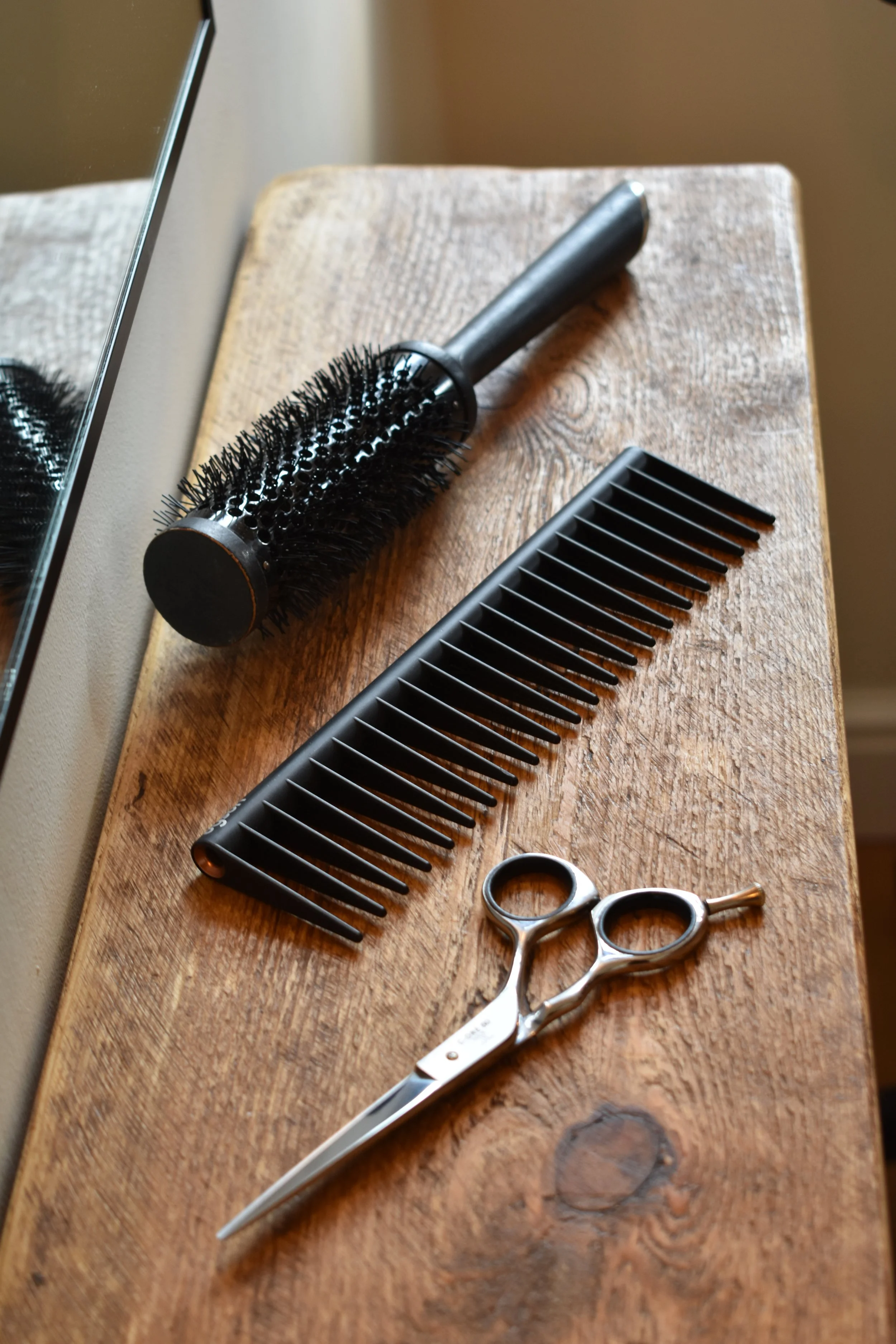A wooden table with a round hairbrush, a wide-tooth comb, and a pair of hair scissors.