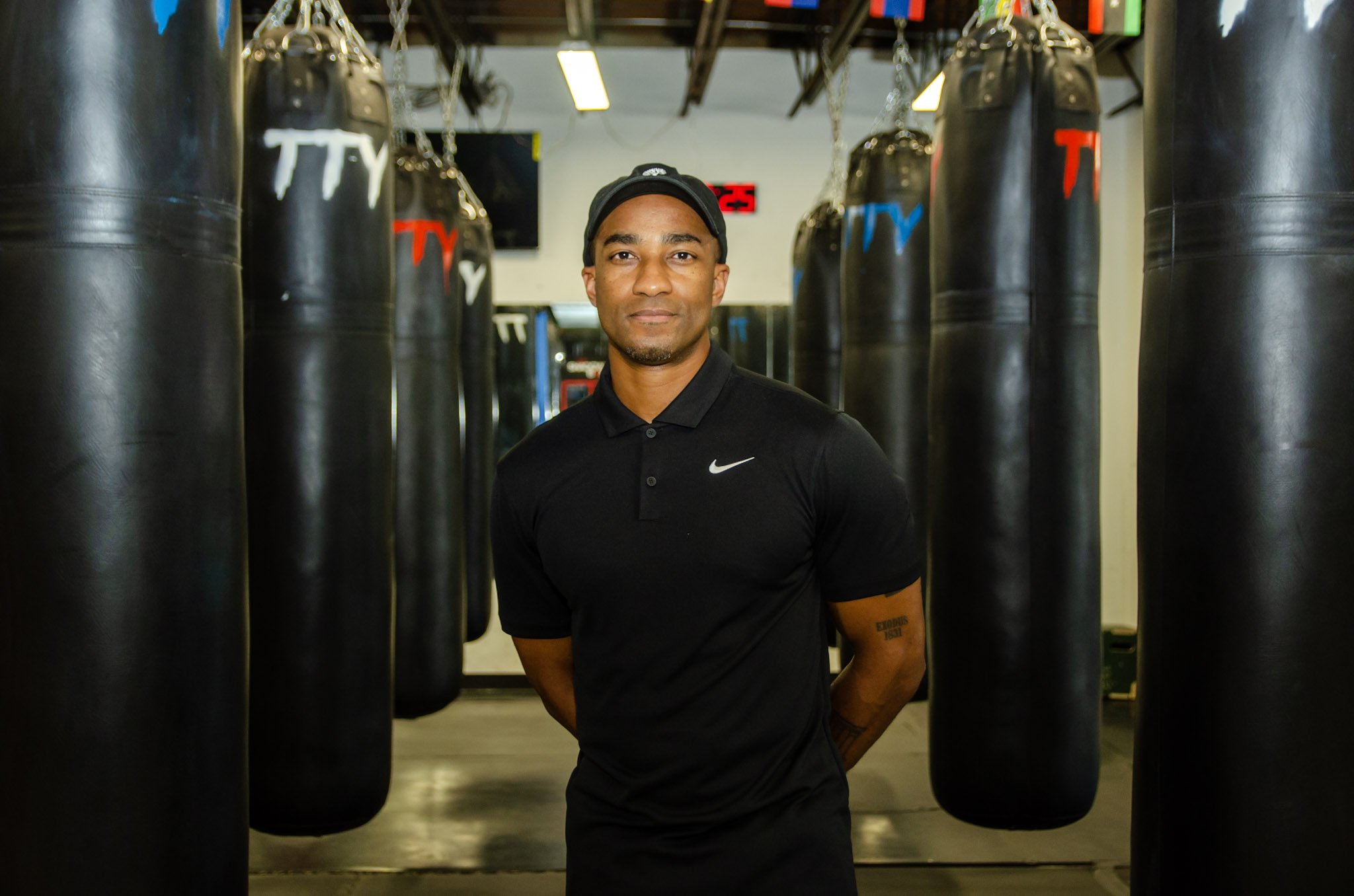 A man standing in a boxing gym surrounded by hanging black punching bags, wearing a black Nike polo shirt and a cap.