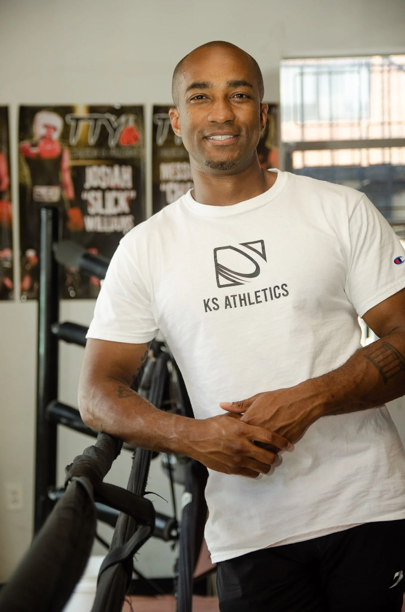 A man smiling, wearing a white KS Athletics t-shirt, standing indoors with workout posters in the background.