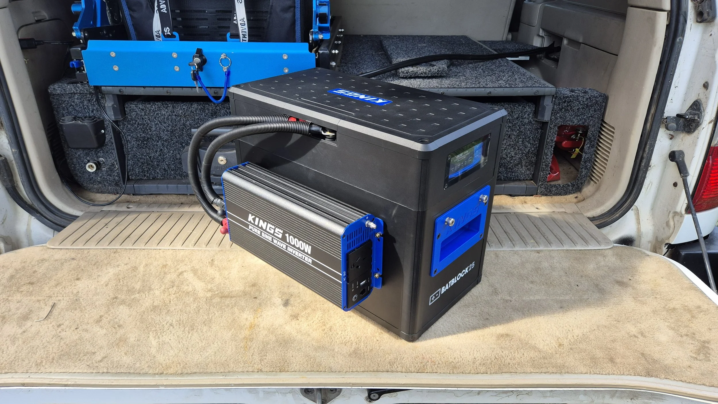 The image shows the interior of a vehicle, possibly an RV or camper van, with electronic equipment installed in the storage area. There is a black and blue inverter labeled "KING'S 1000W" connected with cables, and a blue component with the brand name "BATBLOCK25." The background includes carpeted sections and some wires, with the open rear door of the vehicle visible.
