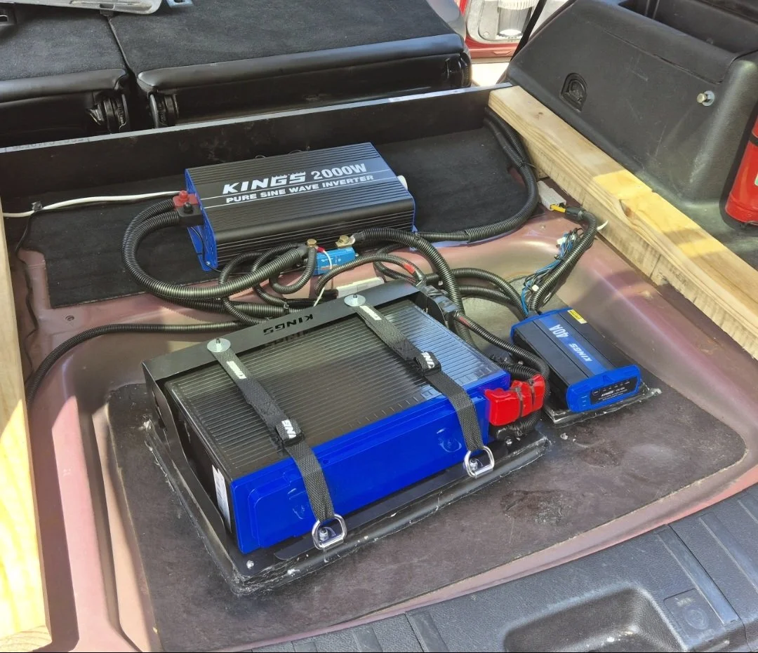 The image shows a setup of power inverters and batteries installed in the trunk of a vehicle, with wiring connecting the devices.