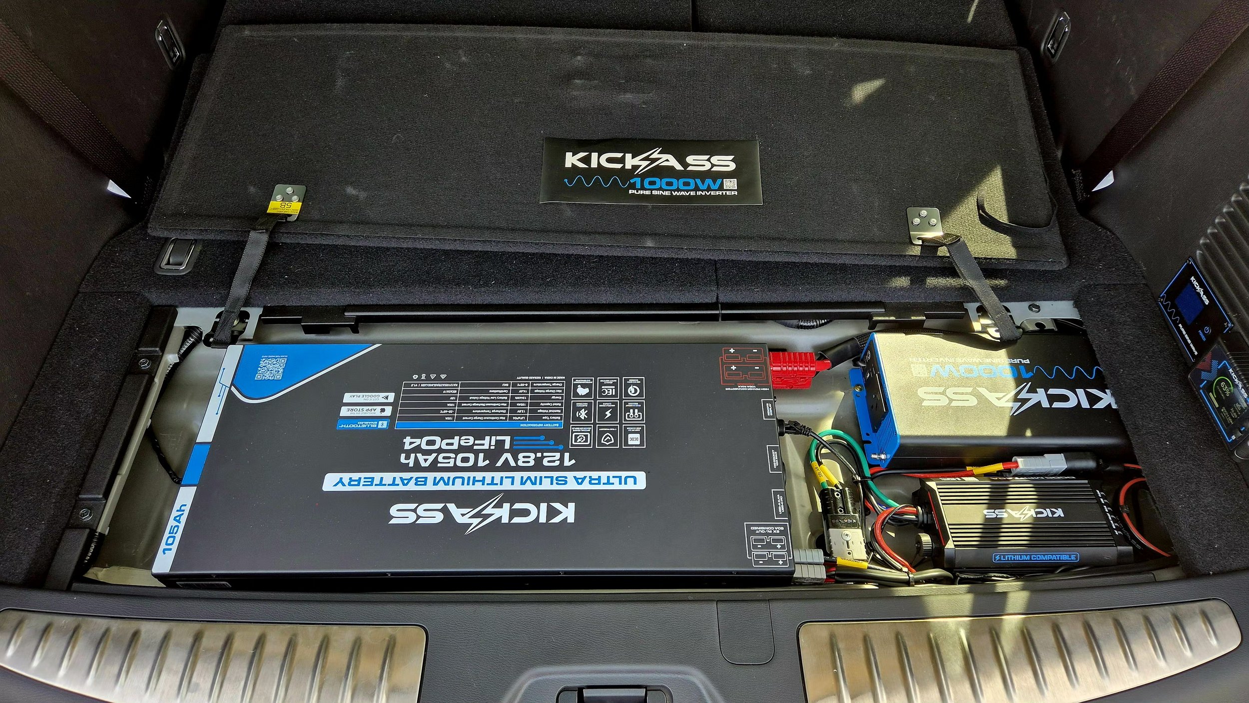 The image shows the inside of a vehicle's trunk with a mounted car audio amplifier, a lithium battery, and a power inverter, arranged neatly.