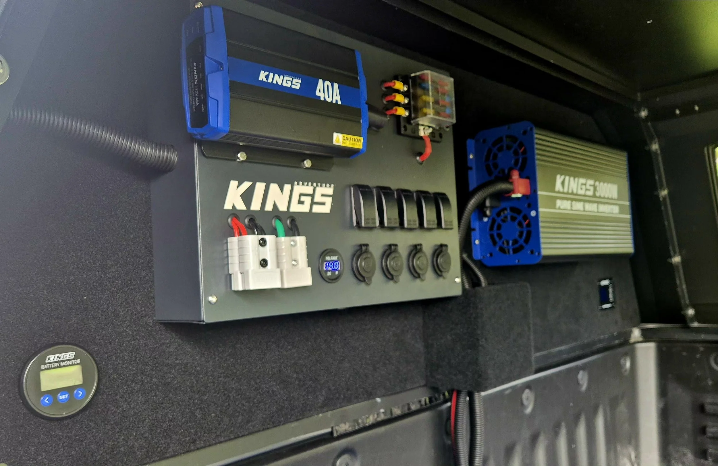 Image of a control panel with various electronic components including a blue Kings 40A battery, a power supply labeled Kings 3000W, a small clear plastic container with colorful wires, a white outlet strip with black and red wires connected, a small digital display, and several switches and knobs. The background shows a dark surface.