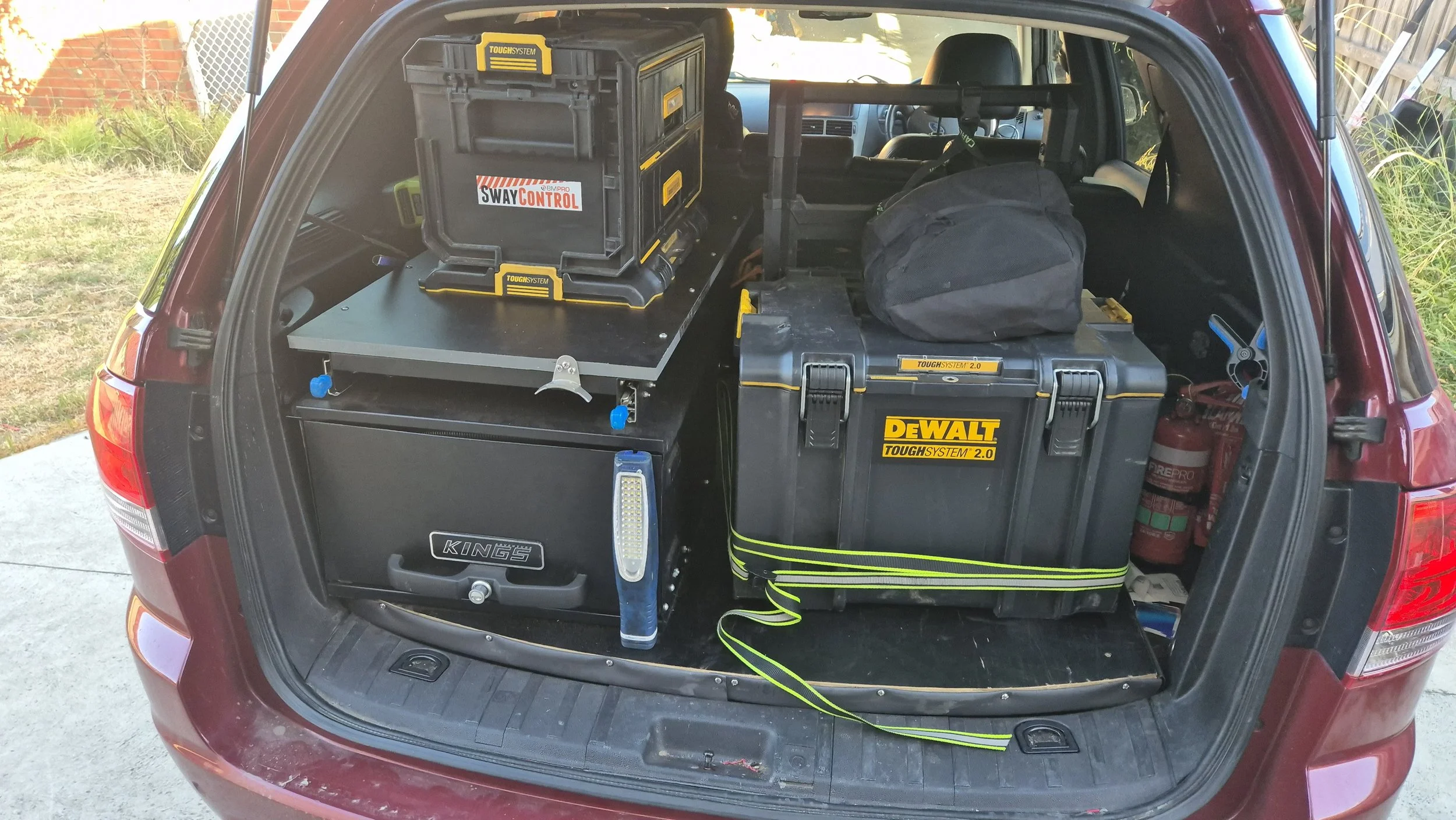 The back of a maroon SUV with the trunk open, showing organized tools and equipment including a large black and yellow Dewalt tough system toolbox, a smaller black Kings toolbox, a black toolbox with a sticker saying Sway Control, a black duffel bag, a fire extinguisher, and some tools and supplies. The vehicle is parked outdoors on concrete, with grass and a fence visible in the background.